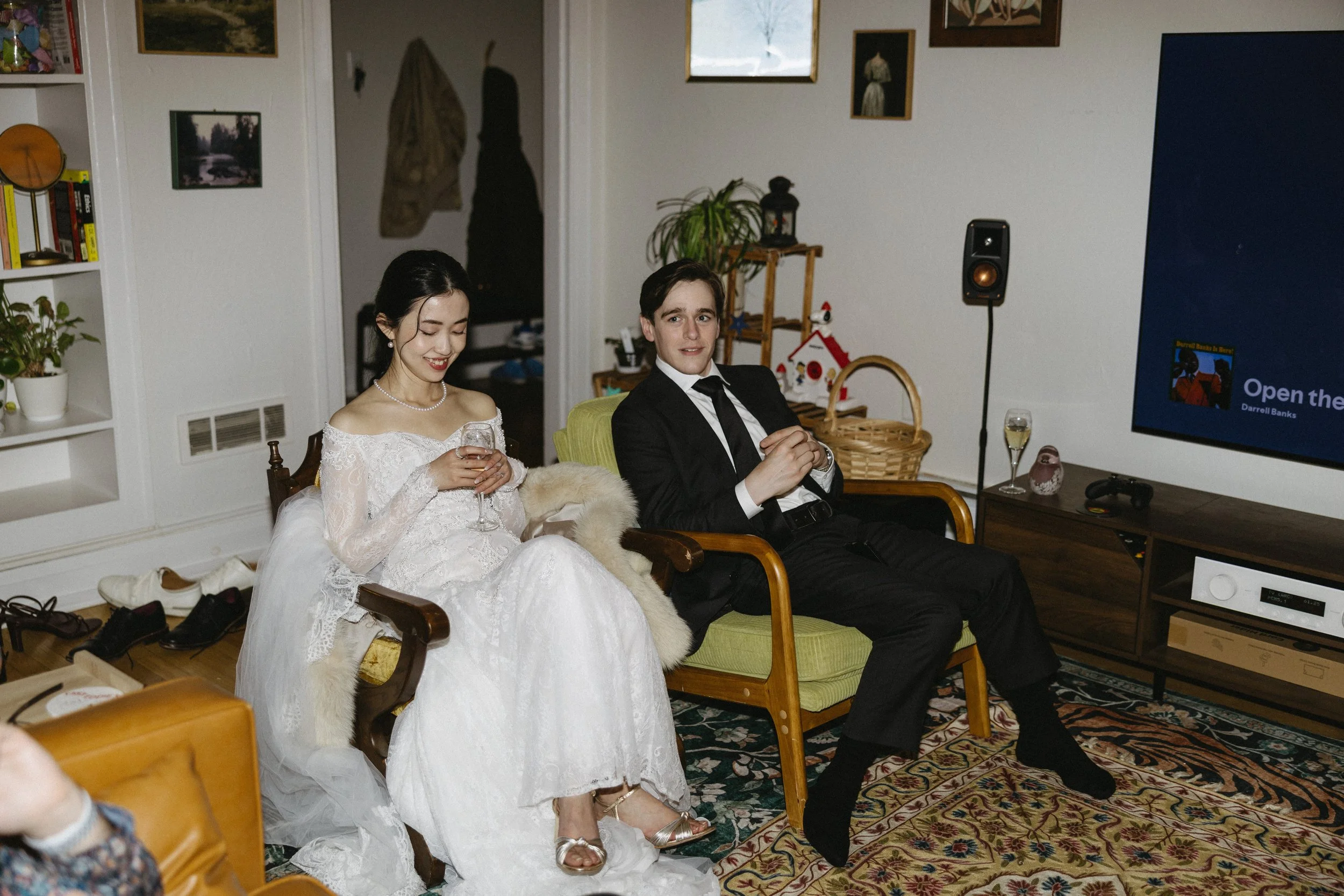 A bride and groom sitting on chairs in a living room during their wedding celebration. The bride is wearing a white lace wedding dress, holding a glass of wine, and smiling. The groom is in a black suit, white shirt, and black tie, sitting with his h