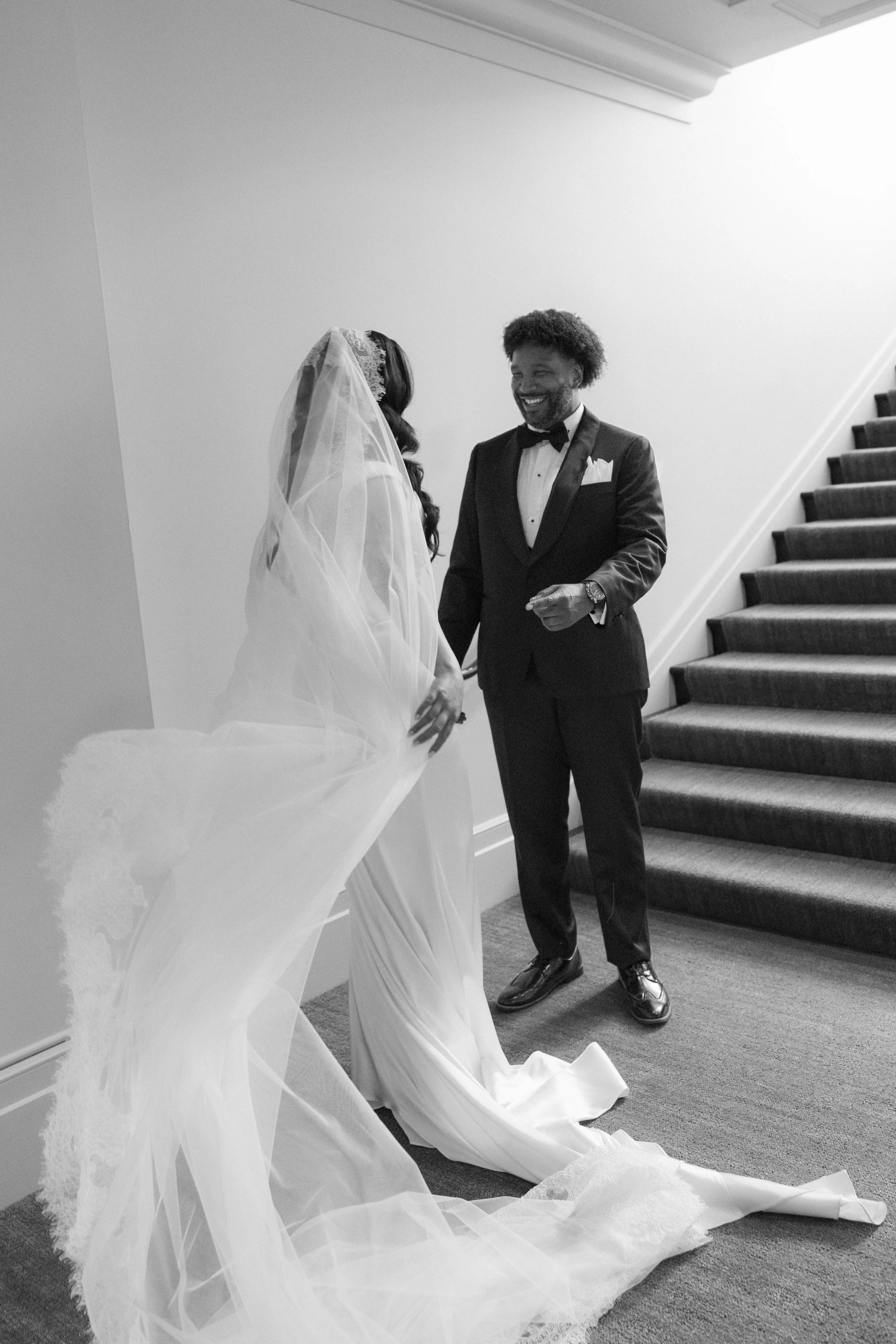 A bride and groom share a joyful moment on their wedding day, standing by a staircase. The bride wears a long, flowing wedding gown with a veil, and the groom is dressed in a tuxedo with a bow tie, smiling at her.