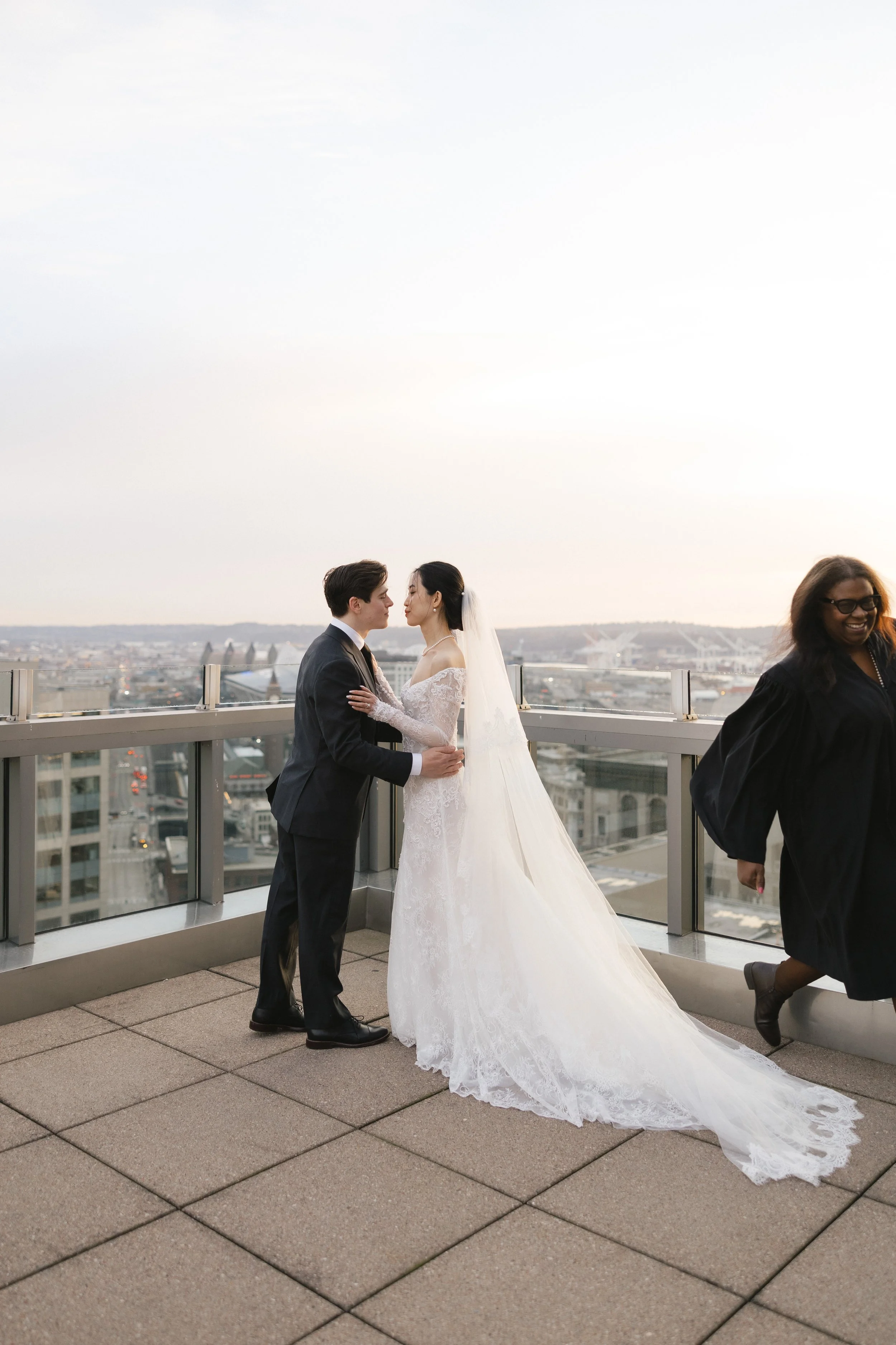 Bride and groom holding hands and facing each other on a rooftop, with a city skyline in the background and a woman in a black gown walking by.