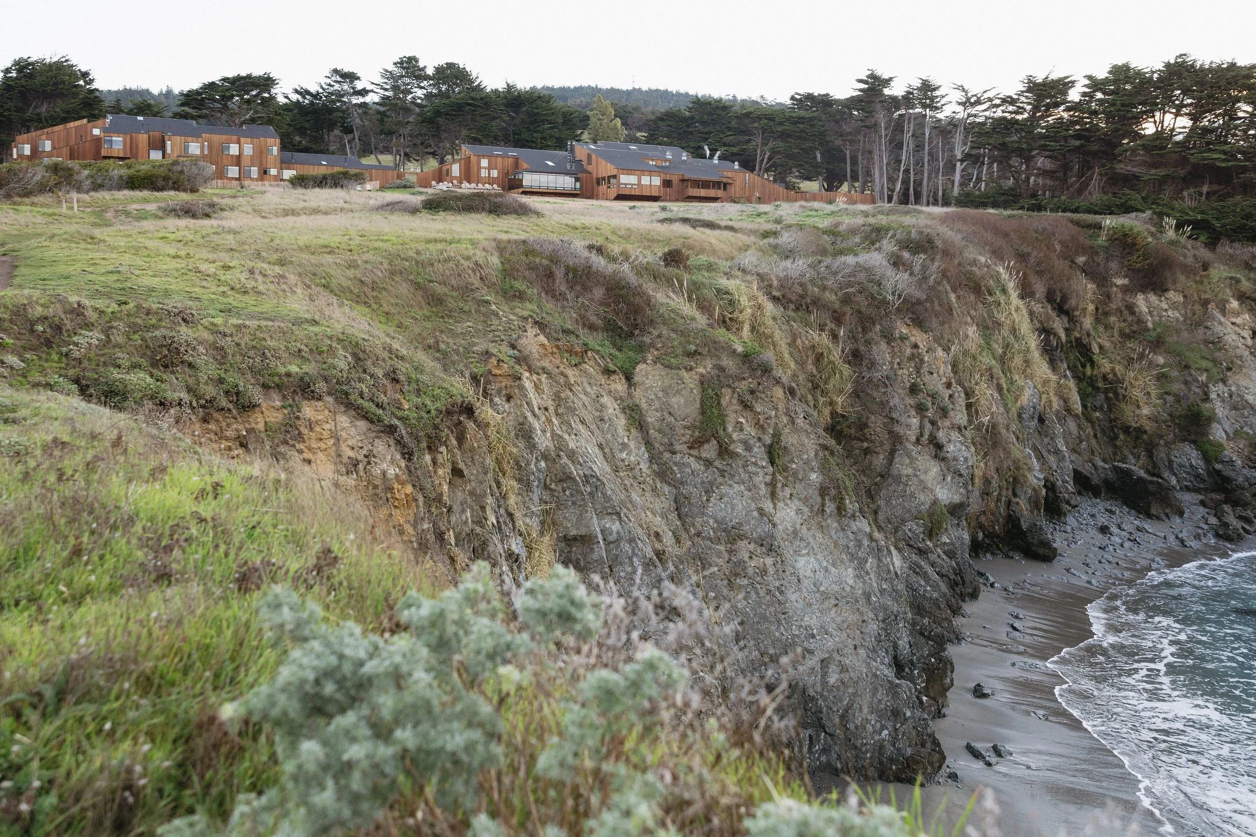 A coastal cliff with a rocky shoreline and grassy top, with modern wooden buildings in the background and trees beyond. Luxury wedding venue in northern california.