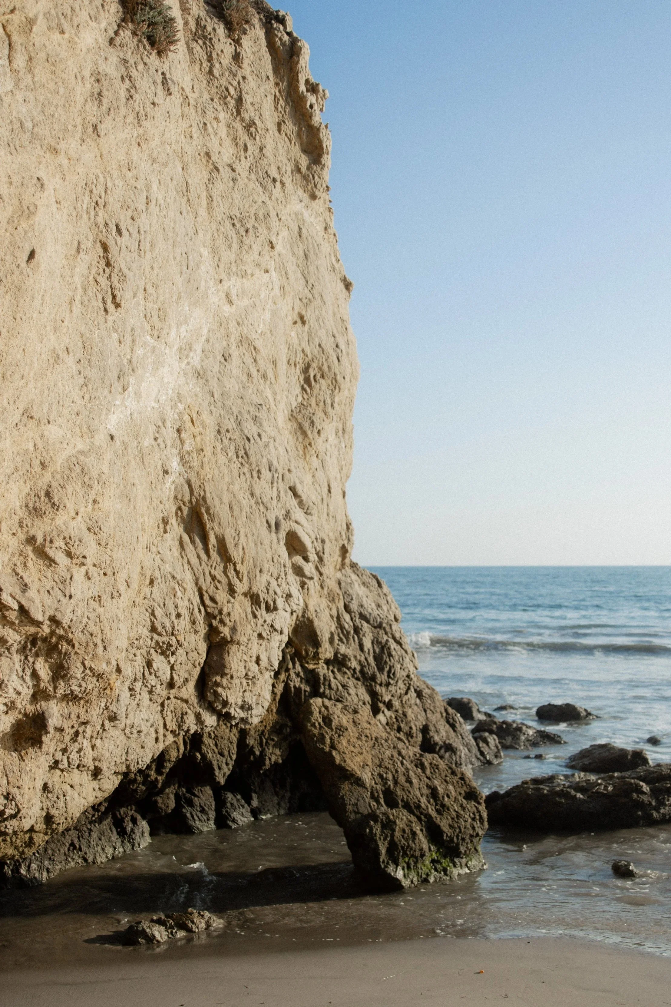 Large sandy sea cliff with rocks at the base, next to the ocean with gentle waves and a clear sky.