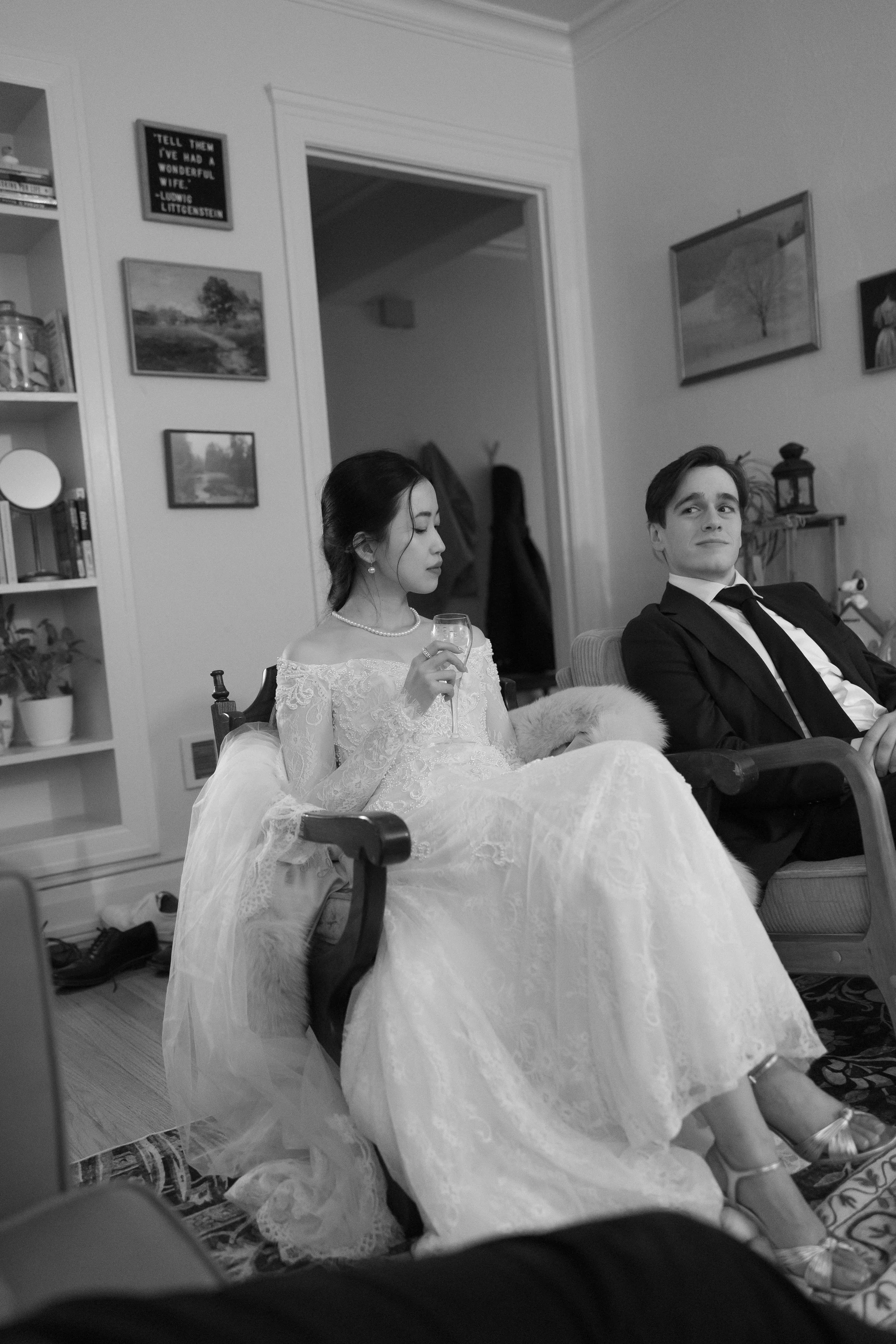 A woman in a wedding dress holding a glass, sitting next to a man in a suit in a living room.