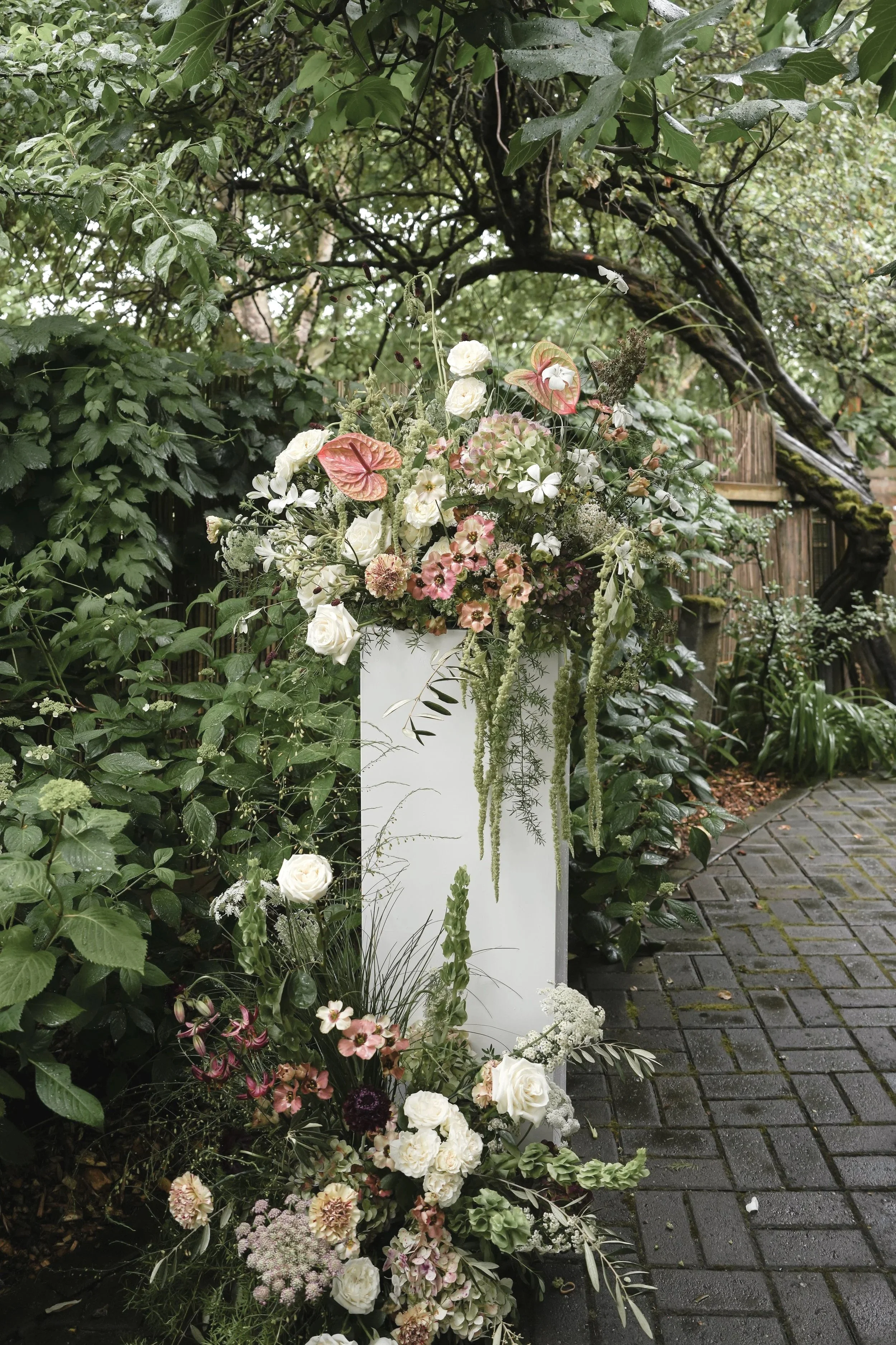 A floral arrangement on a white stand with various white, pink, and green flowers, set outdoors along a cobblestone path, surrounded by lush green foliage and trees. the corson building wedding venue.