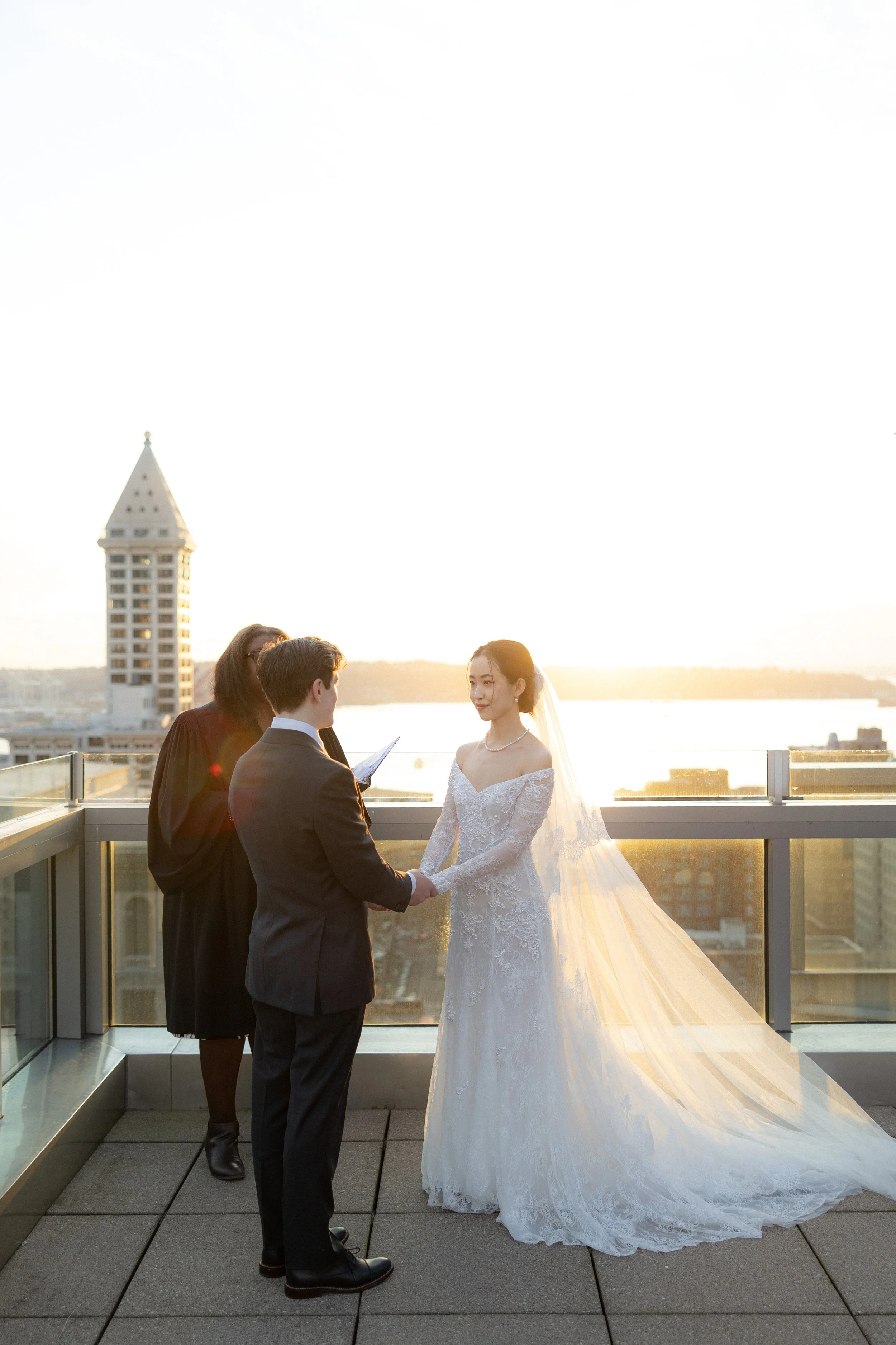 A wedding ceremony on a rooftop during sunset with a bride in a lace gown and veil, a groom in a suit, and two officiants, overlooking a cityscape and water.