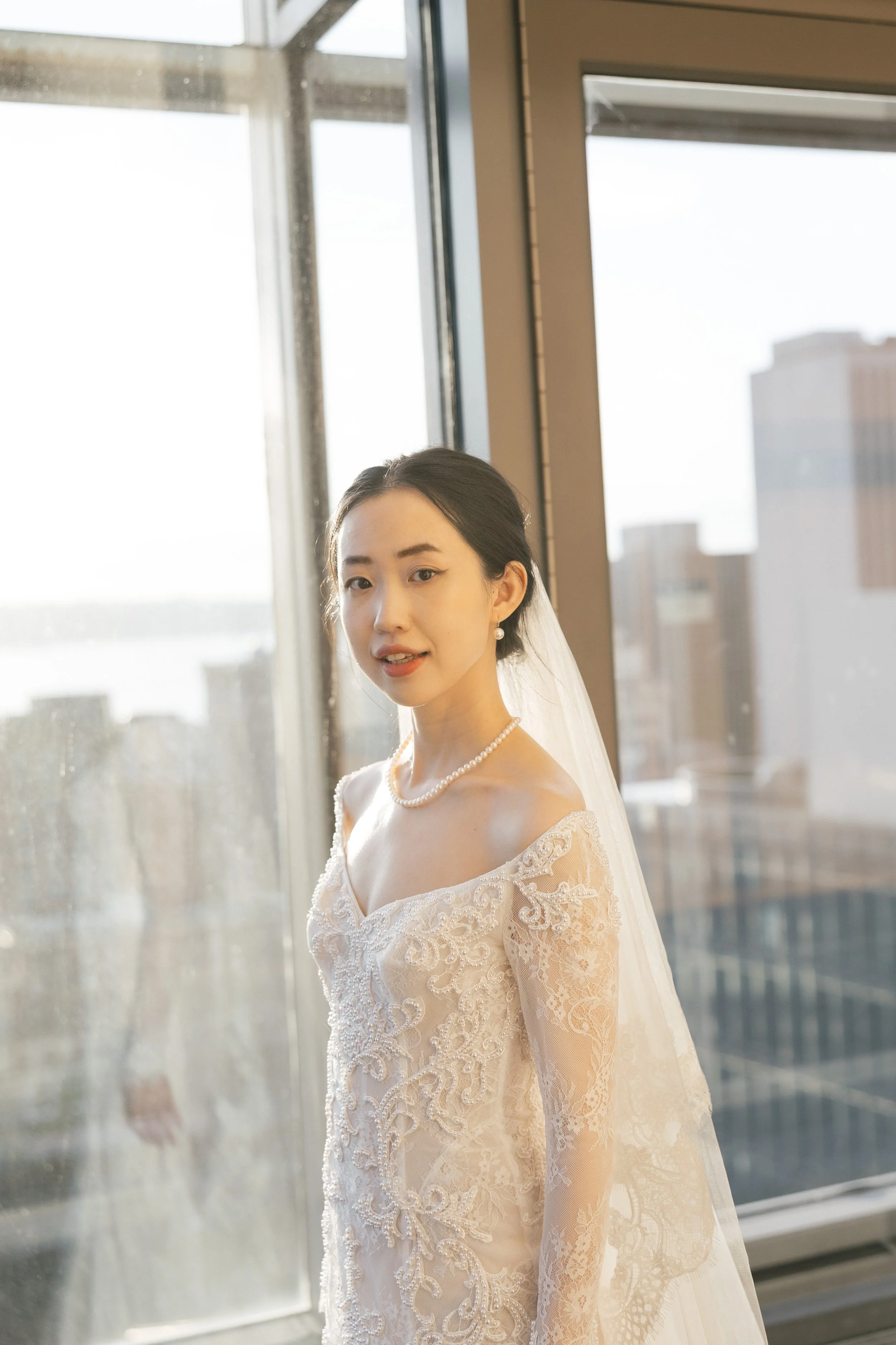 A bride in a lace wedding dress and pearl jewelry standing near a large window with city buildings and water in the background.