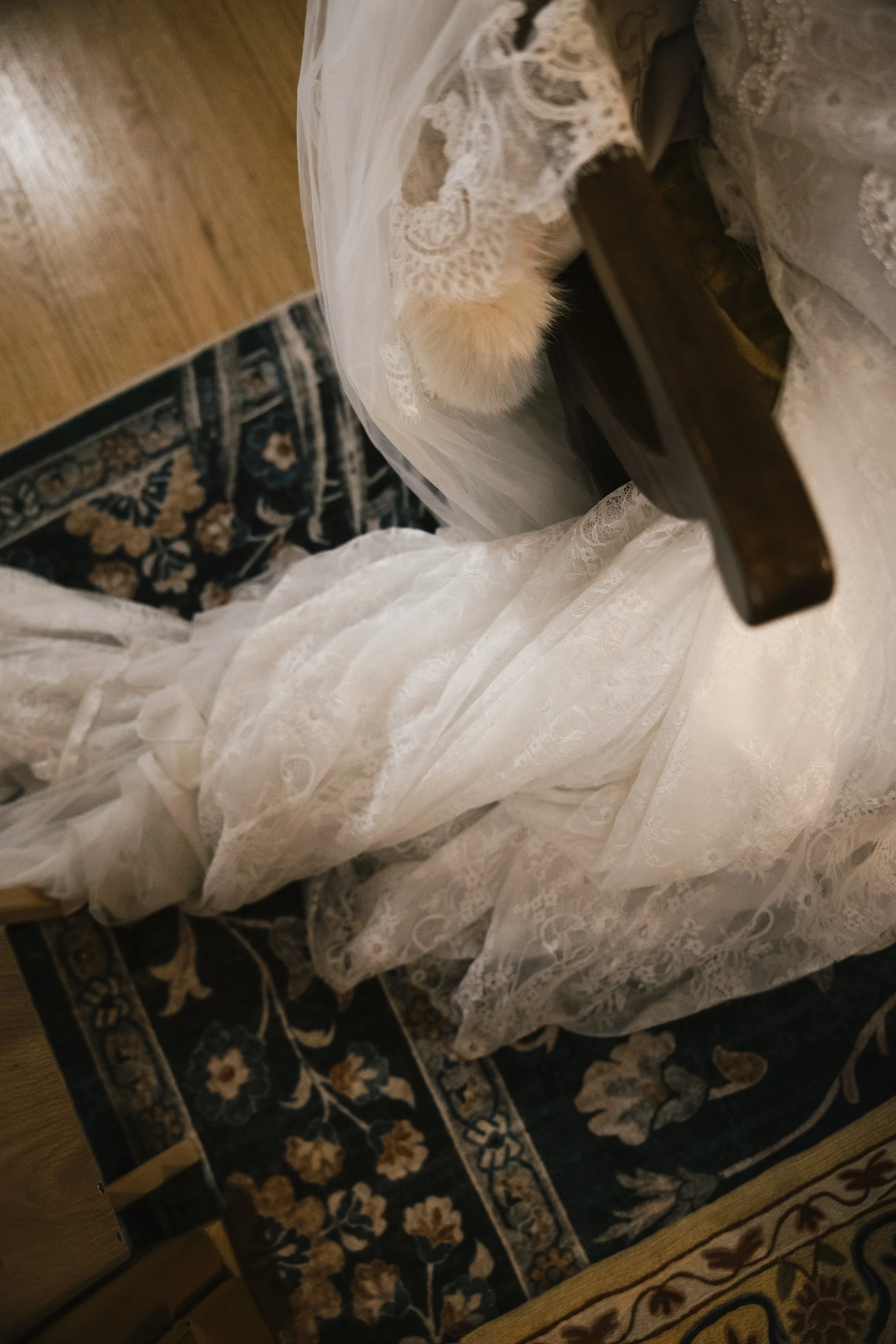 A wedding dress with lace details and a long train, hanging from a piece of furniture, seen from above. Part of a patterned rug and wooden floor are also visible.