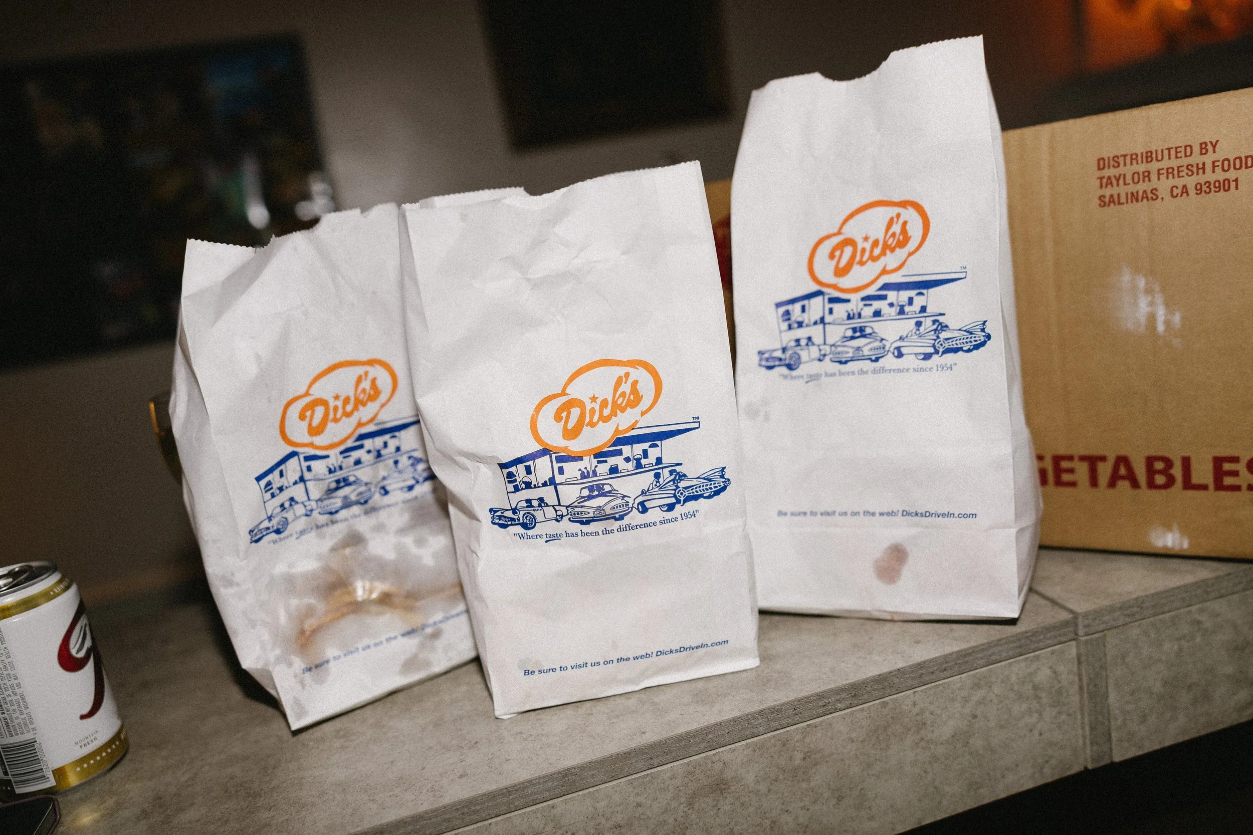 Three crumpled paper bags with Dick's logo and drawings of cars, placed on a gray countertop with a can of soda nearby and a cardboard box labeled 'vegetables' in the background.