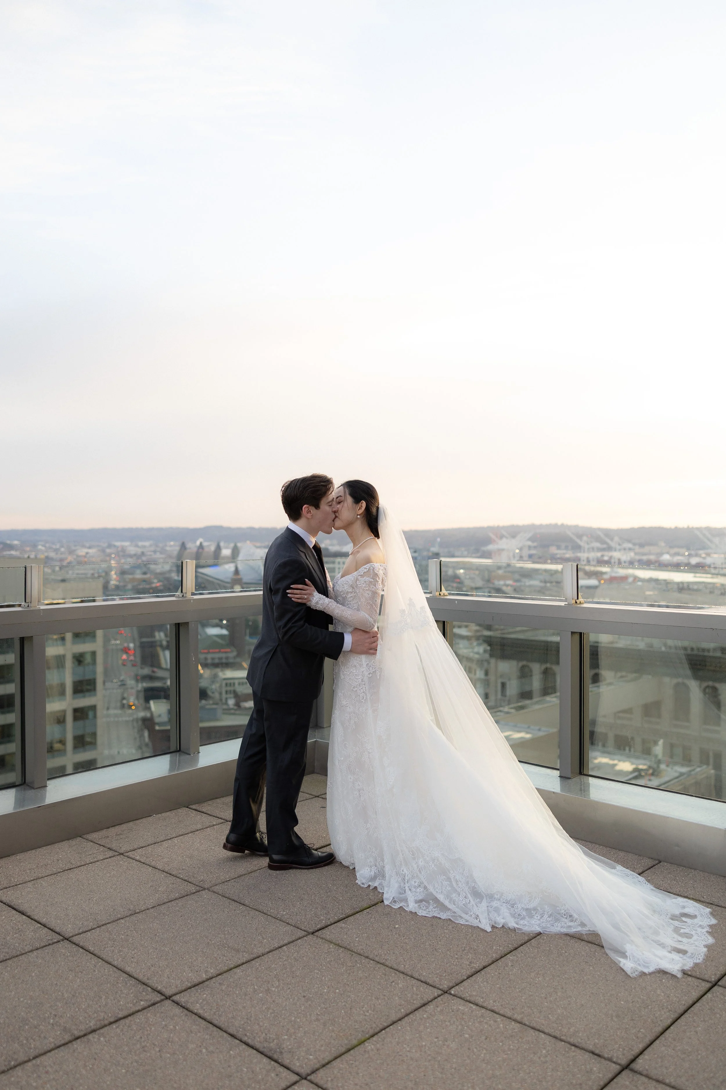 A bride and groom sharing a kiss on a rooftop balcony during sunset, with a city skyline in the background.