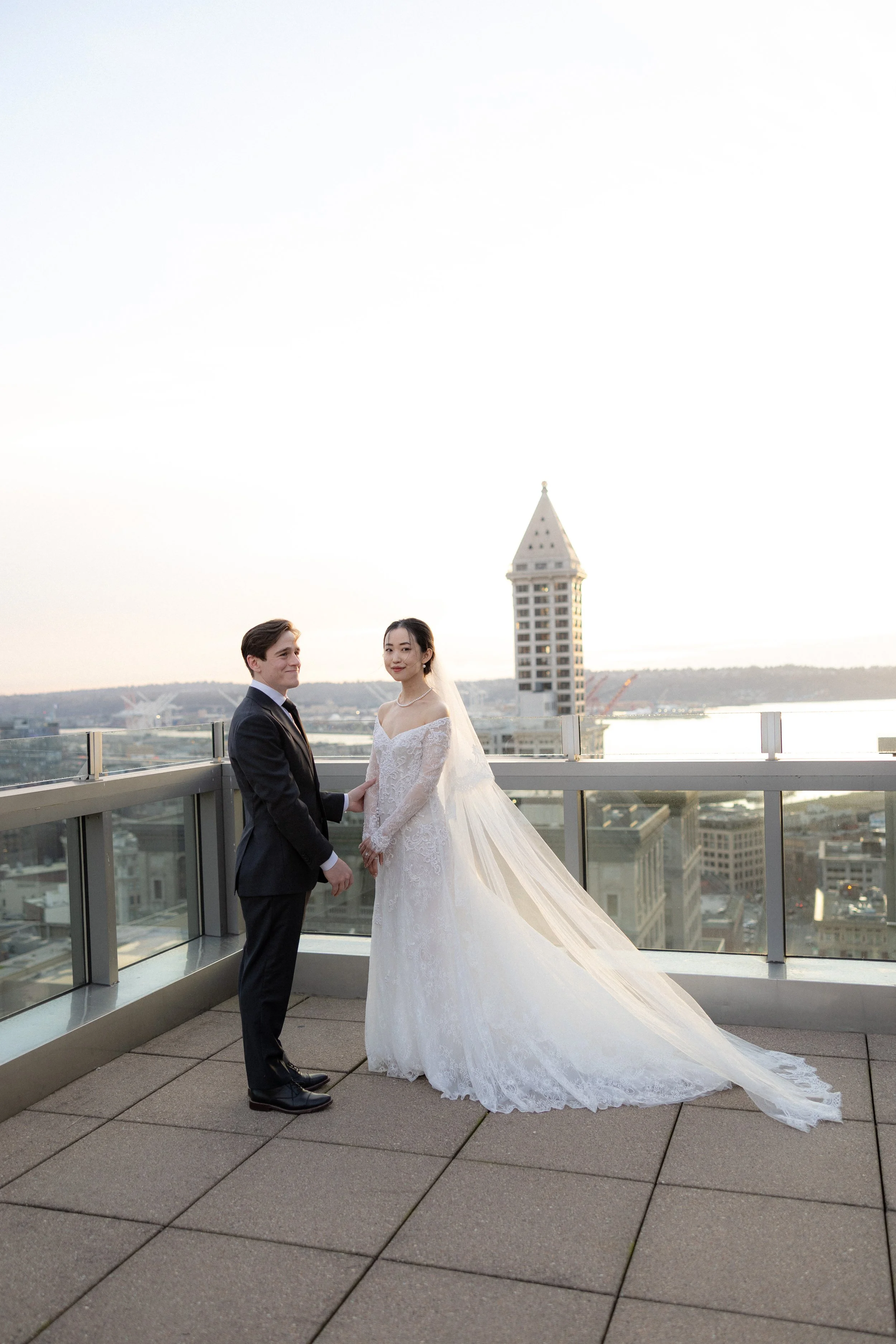 A bride in a white lace wedding gown with a long train and veil holding hands with a groom in a black suit on a rooftop with city view and a tall building in the background.