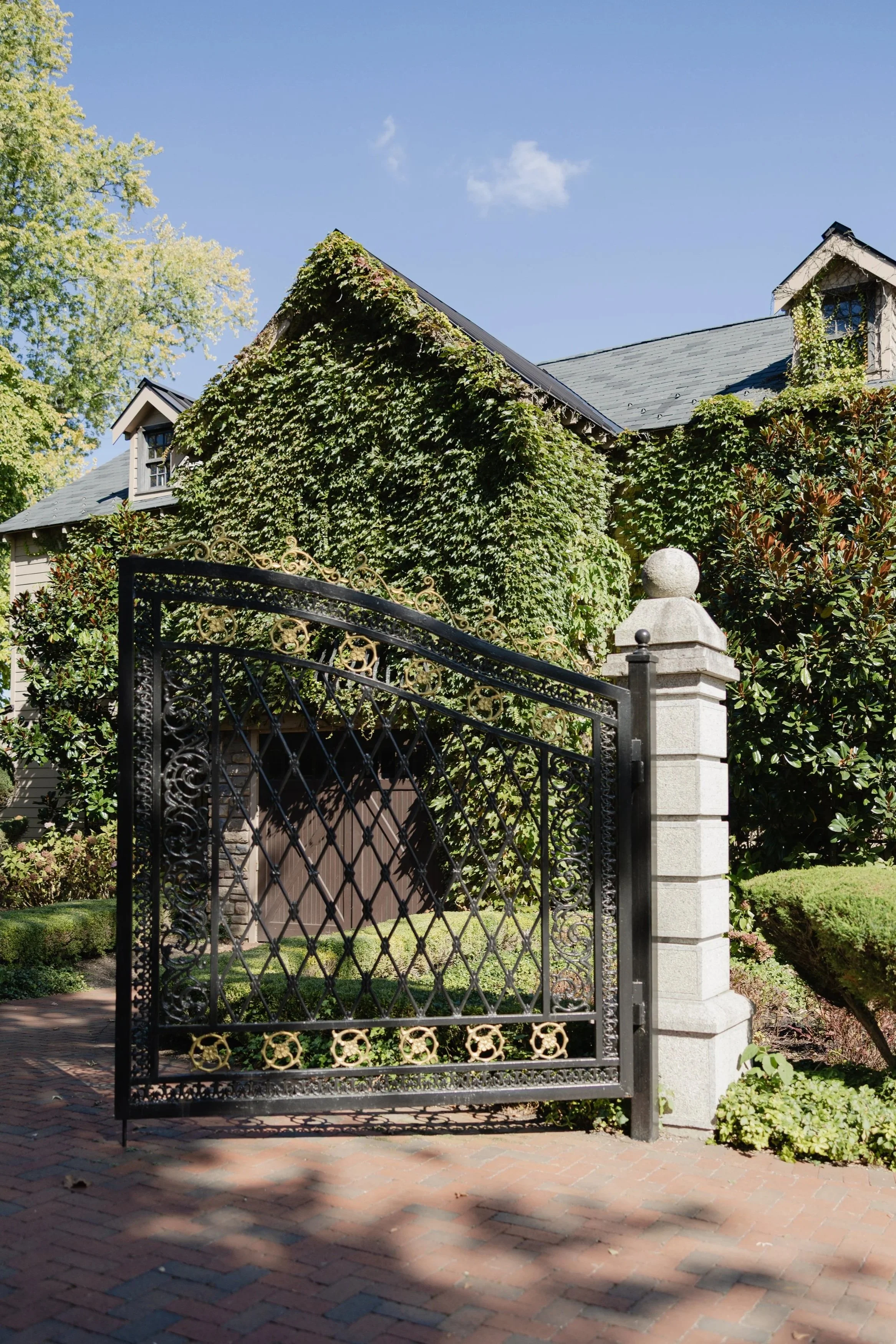 A house with ivy growing on the front wall, a black wrought-iron gate, and a stone pillar, with a brick pathway and lush green bushes and trees.