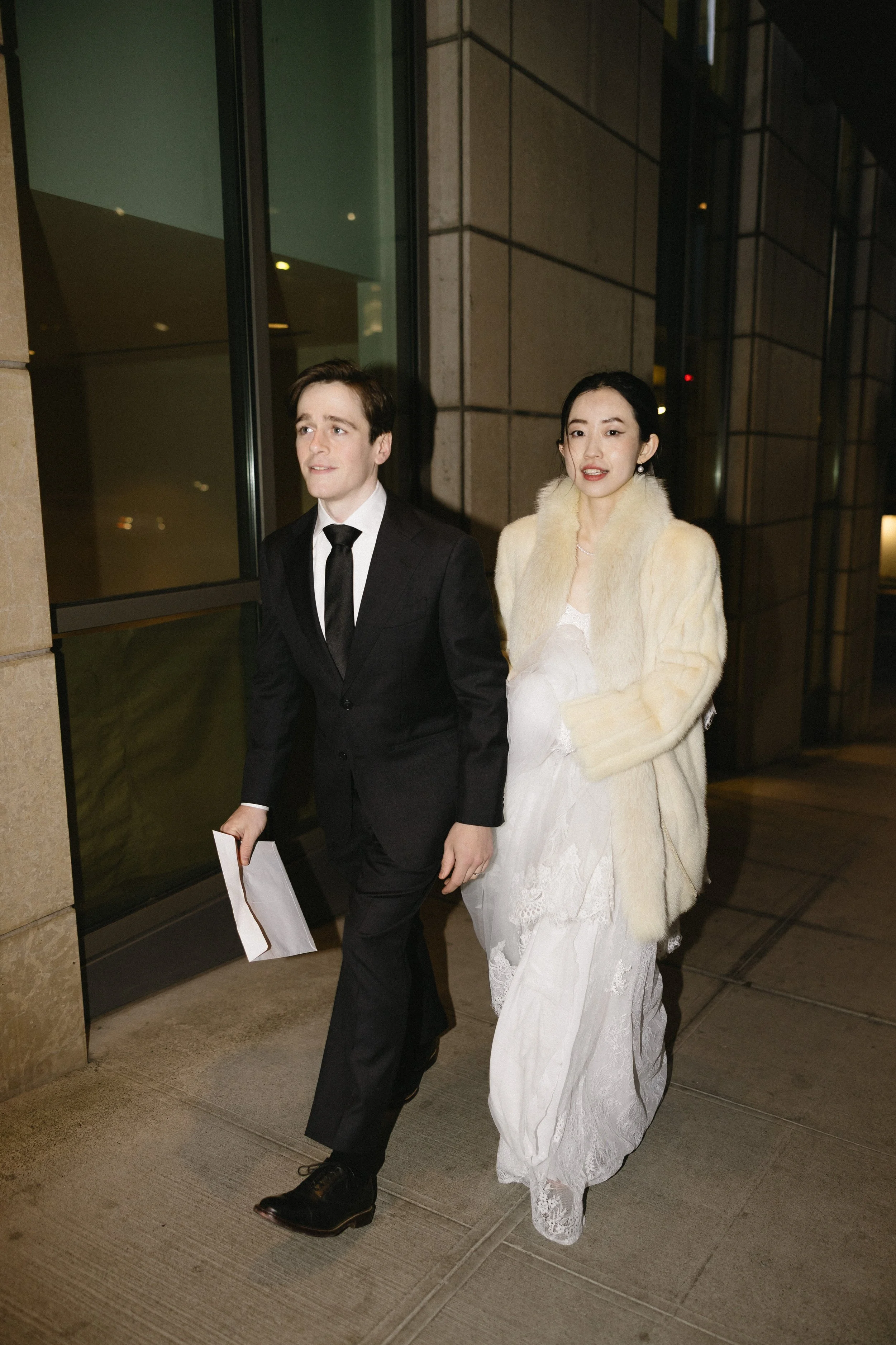 A man in a black suit and tie walking beside a woman in a white dress and cream-colored fur coat outside a building at night.
