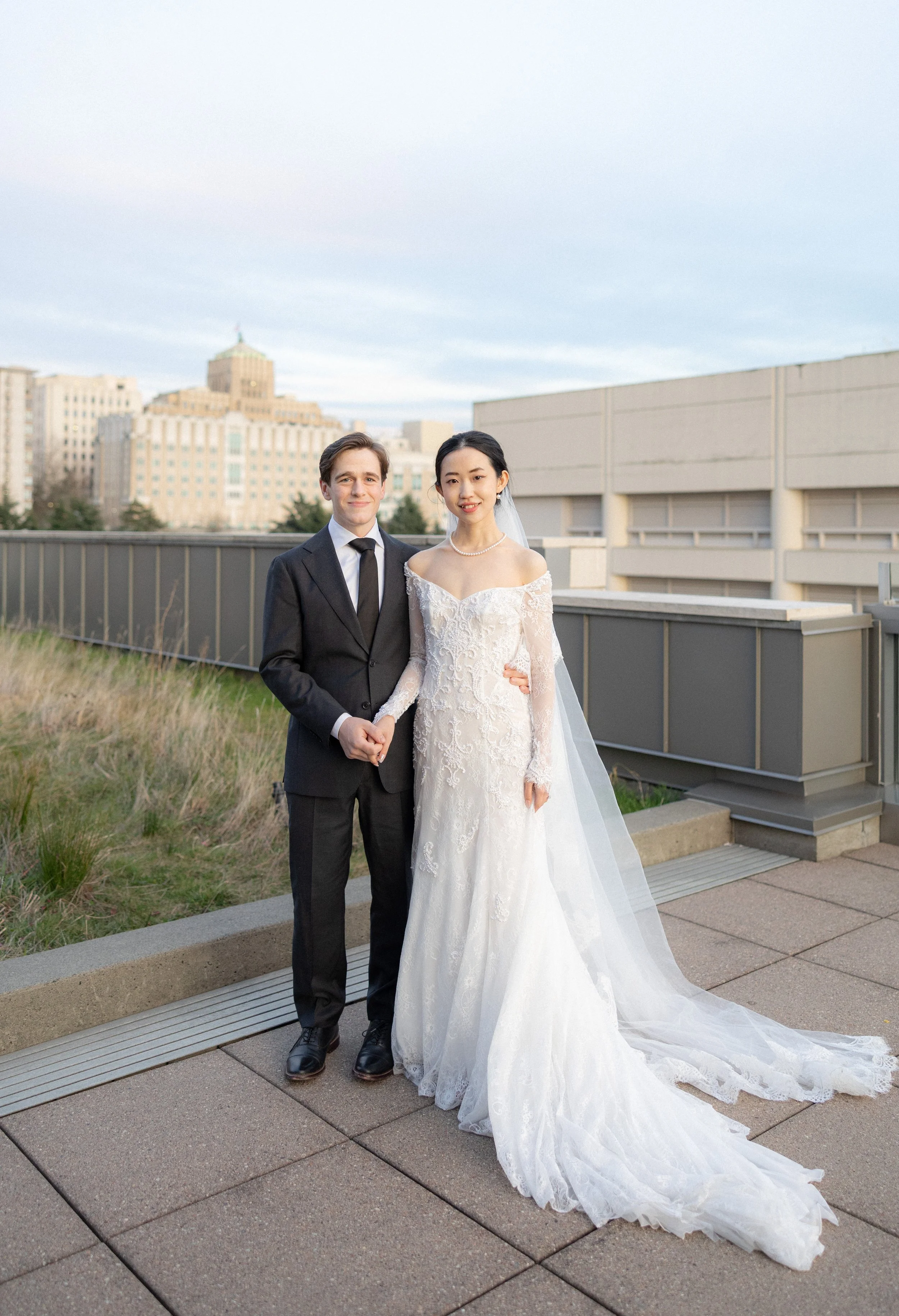 A newlywed couple holds hands on a rooftop with a city skyline in the background. The bride wears a white lace wedding gown with an off-the-shoulder neckline and a long train, and the groom wears a black suit with a white shirt and black tie.