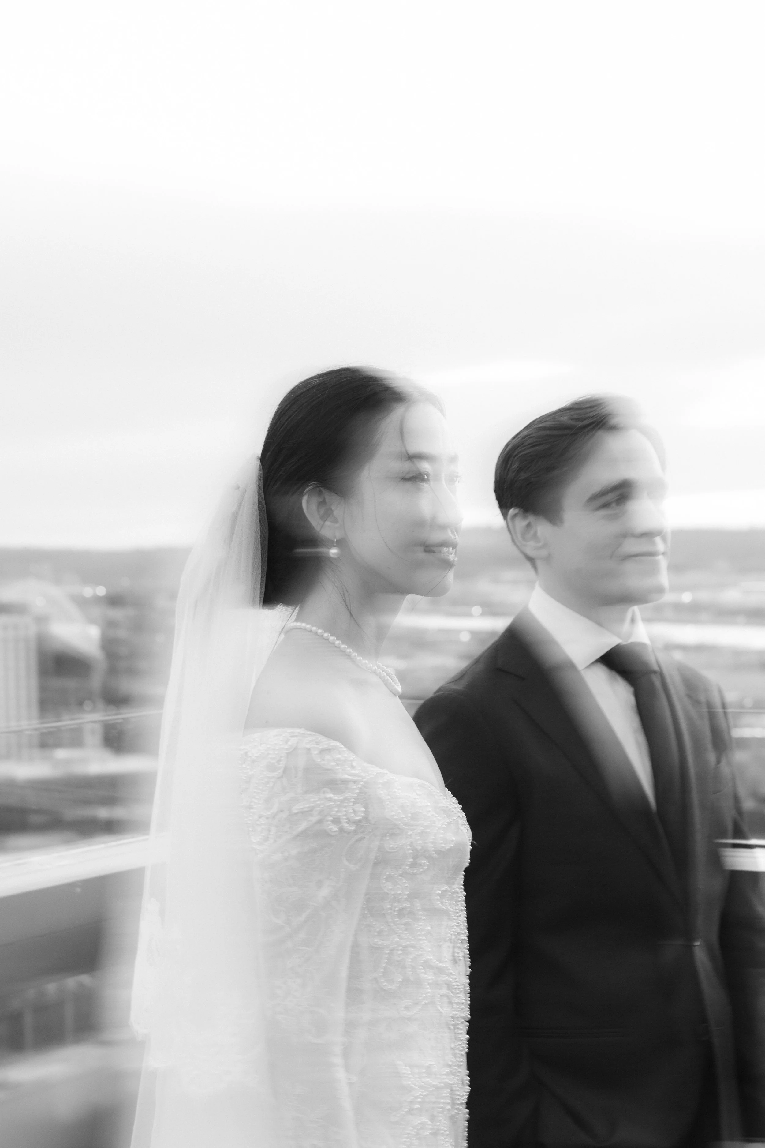 Black and white photo of a bride and groom standing side by side on a balcony, with cityscape in the background, a wedding scene.