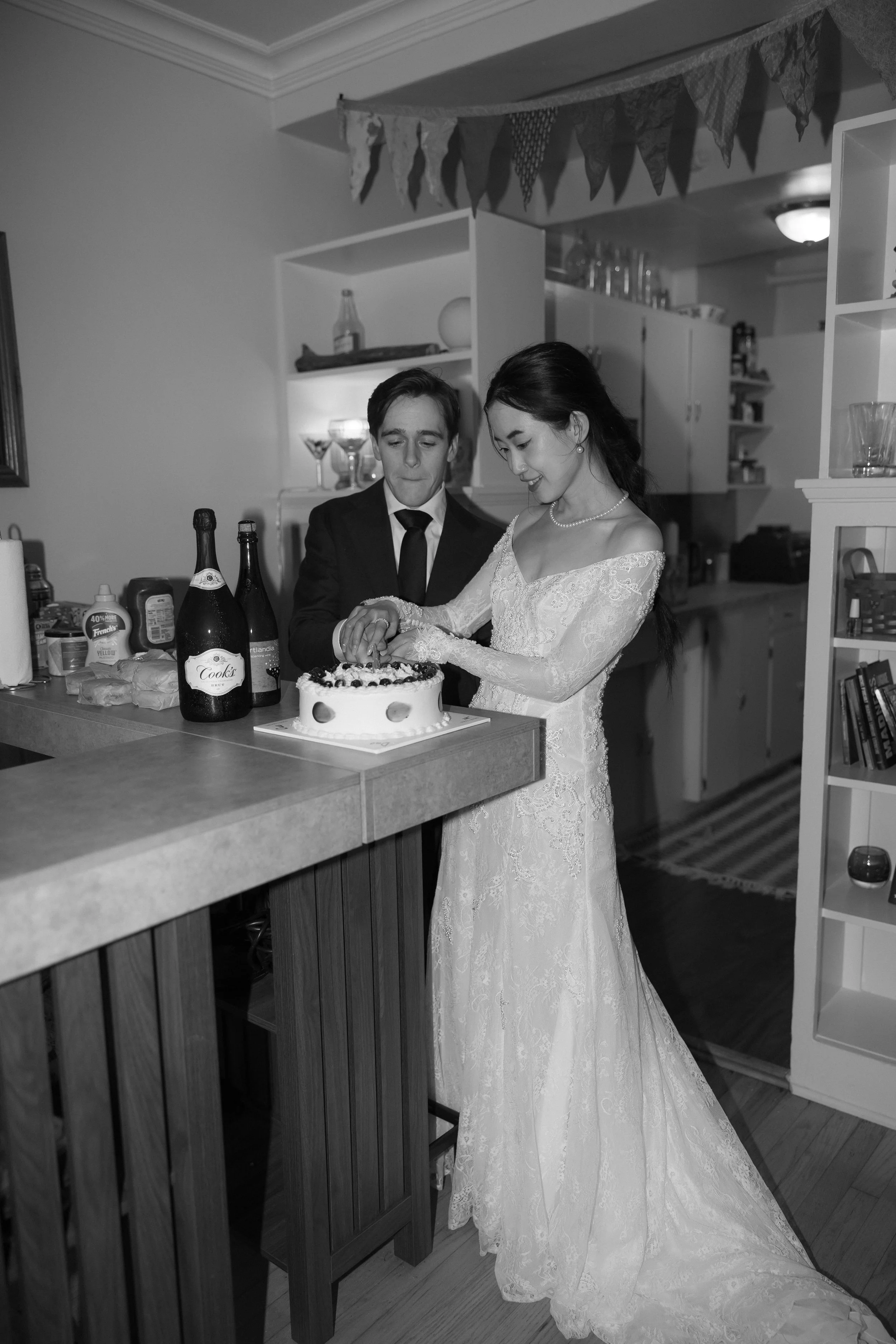 A bride in a lace wedding dress and a groom in a suit cut a cake together in a kitchen decorated for a celebration.