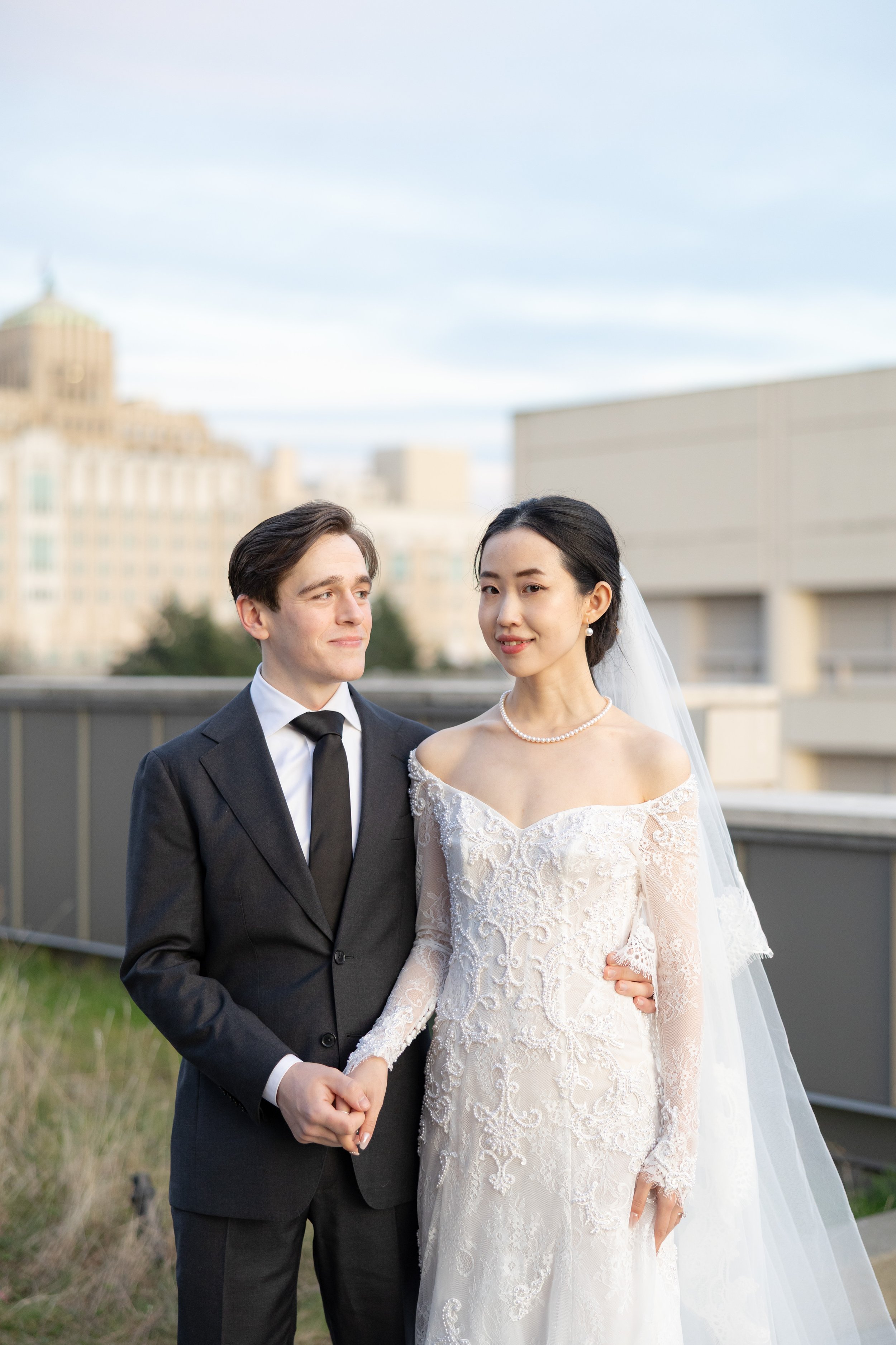 A bride and groom hold hands outdoors with city buildings in the background, the bride in a lace wedding dress and pearl necklace, the groom in a dark suit and tie.