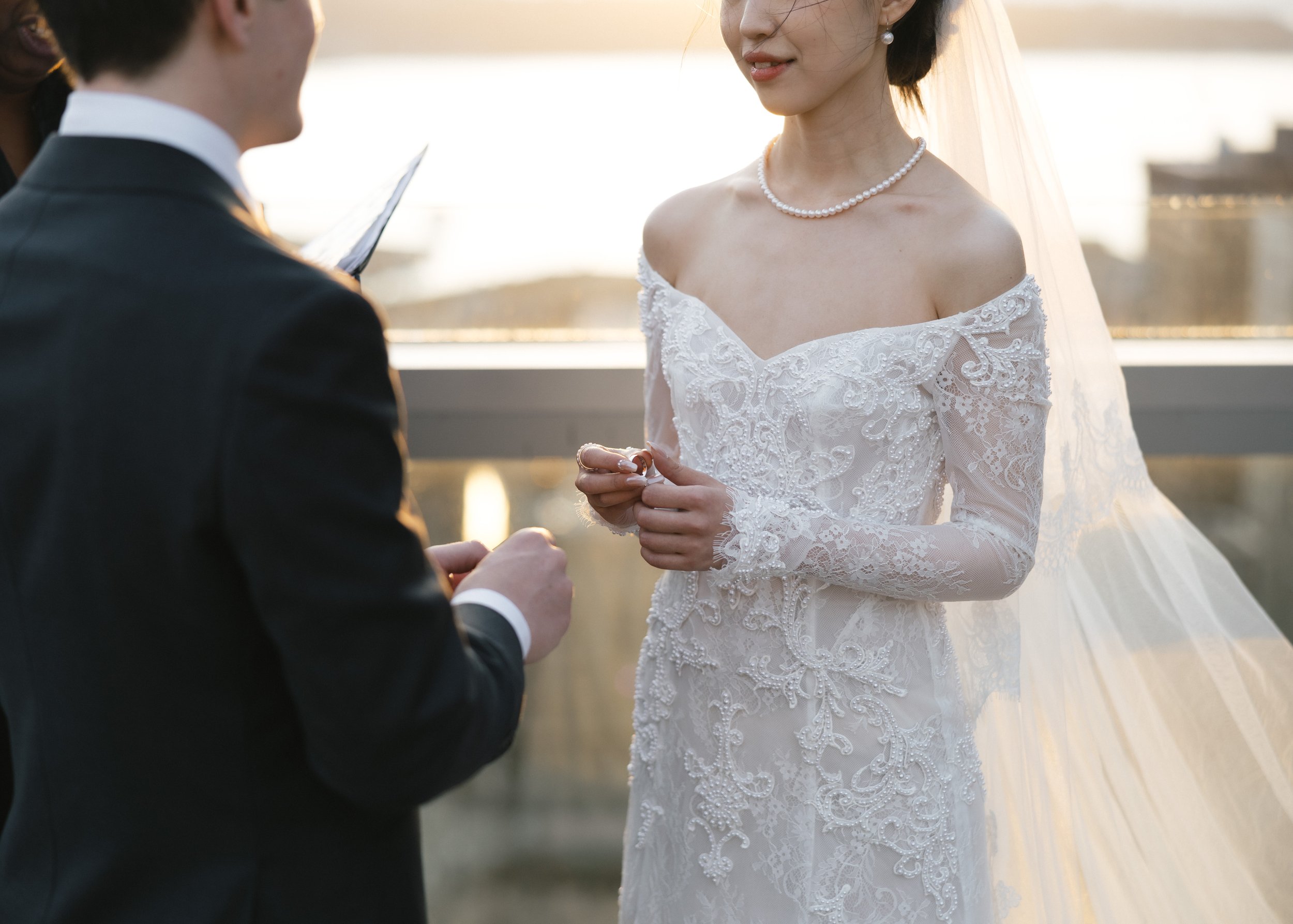 A bride and groom exchanging rings during their wedding ceremony outdoors at sunset.