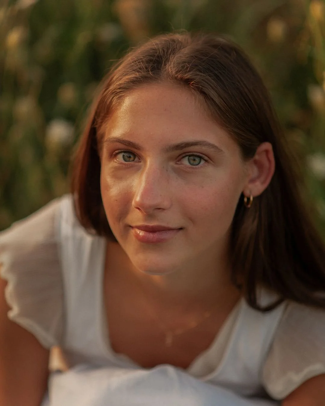 Close-up of a young woman with blue eyes and brown hair, wearing a white top and small hoop earrings, outdoors with blurred greenery in the background.
