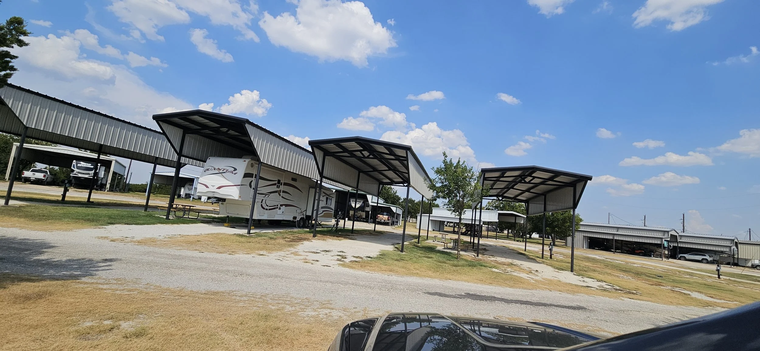 Carport structures sheltering several RVs and vehicles under a partly cloudy sky with a few trees in the background.