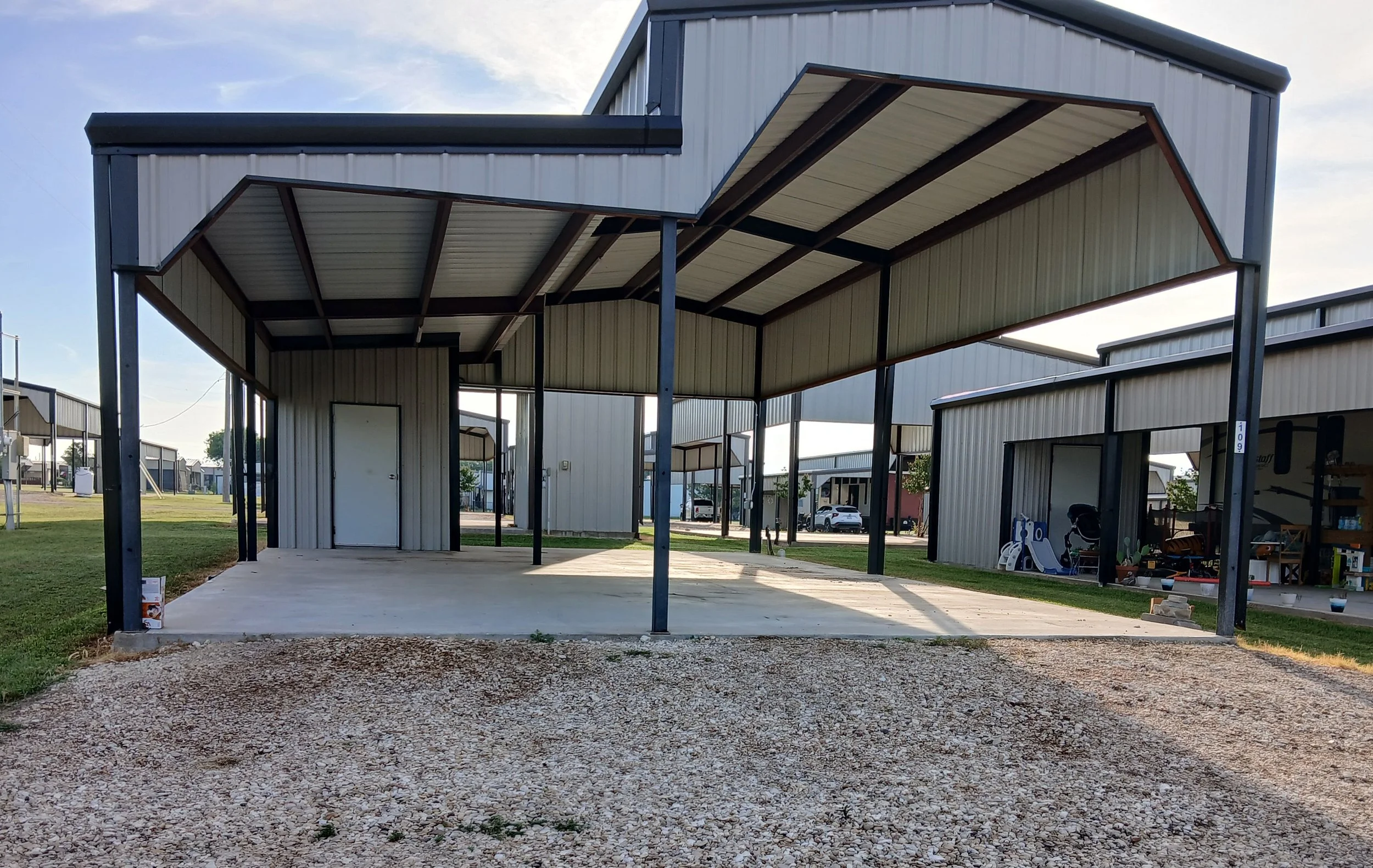 Metal carport with a beige roof and black support beams in a residential area, with a concrete slab floor and surrounding grass and gravel.