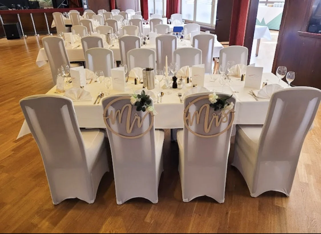 Ein festlich gedeckter Hochzeitstisch mit weißen Stuhlhussen, zwei Stühlen vorne mit "Mr" und "Mrs" Schildern, Blumenarrangements, Wein-, Wasser- und Weingläser, sowie Kerzen und Menükarten.