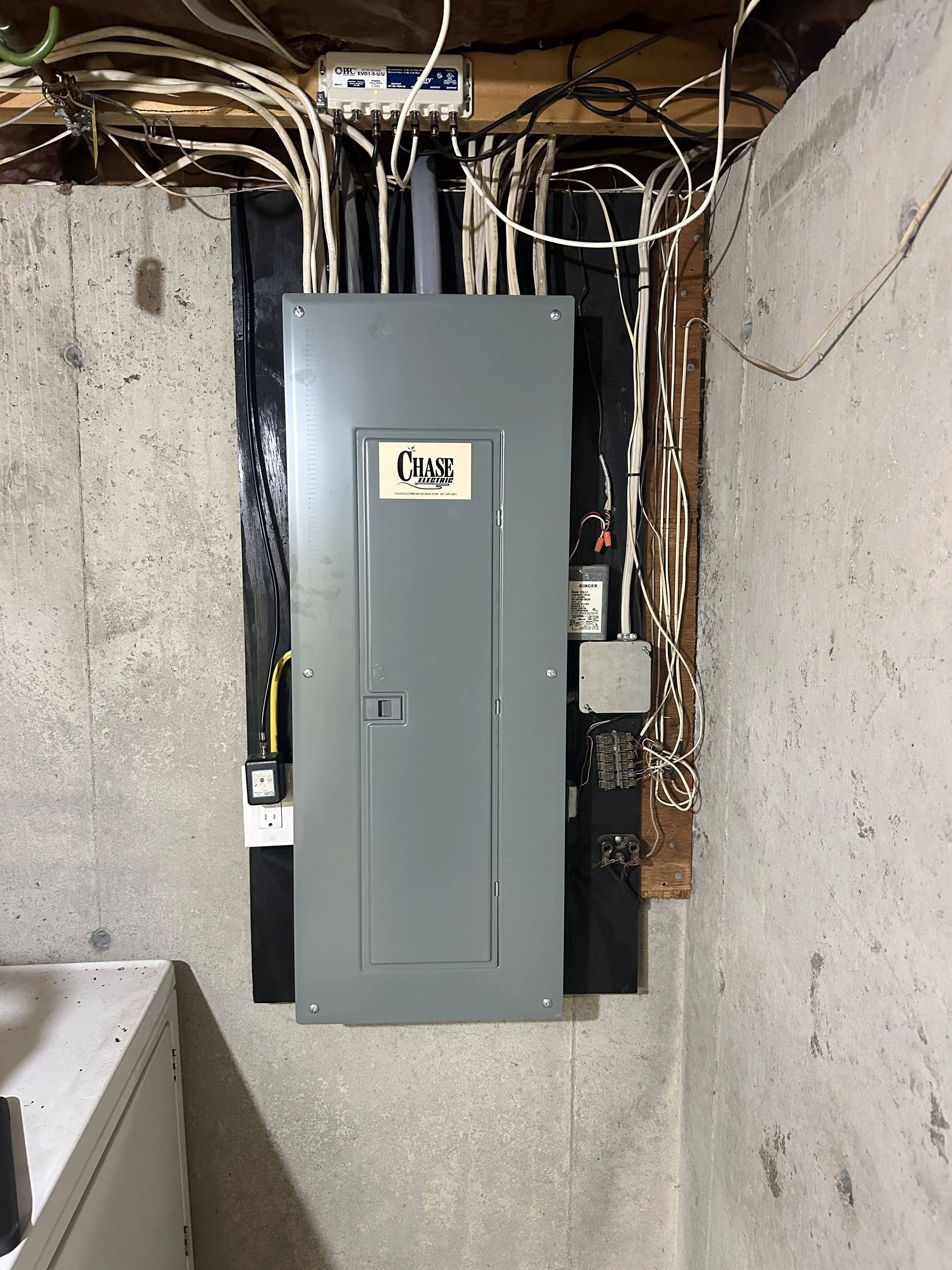 Electrical panel and wiring mounted on a concrete wall, with multiple white and black wires connected to it.