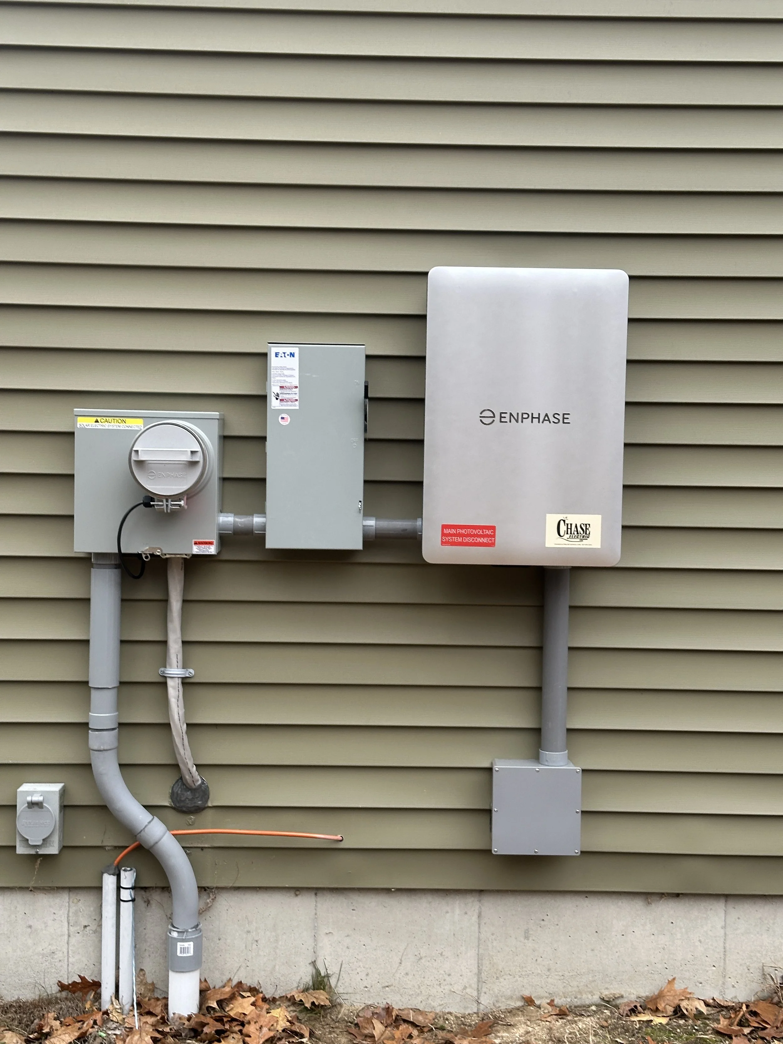 Outdoor electrical and solar equipment mounted on a house wall, including an Enphase solar inverter, electrical boxes, and wiring with leaves on the ground.