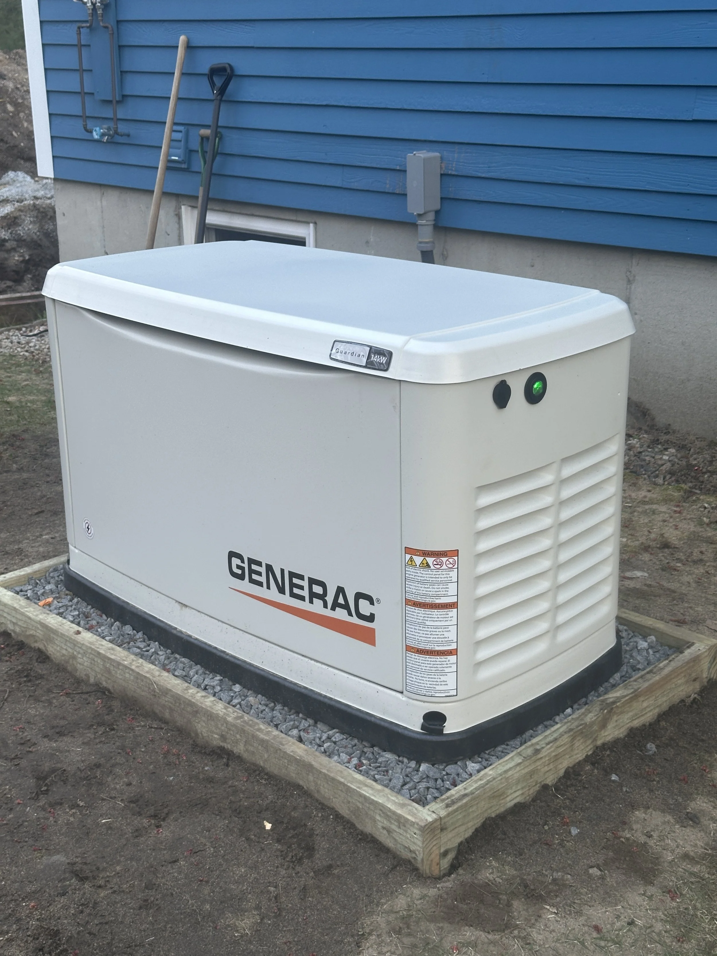 A Generac home standby generator installed on a wooden platform next to a house with blue siding, with shovels leaning against the house.