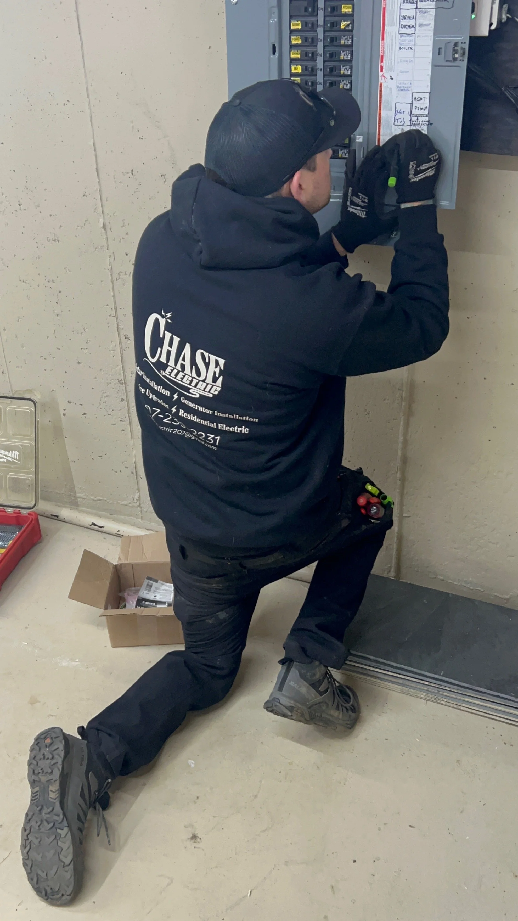 A utility worker kneeling on one knee, wearing a black hoodie and gloves, working on an electrical panel on a beige wall.
