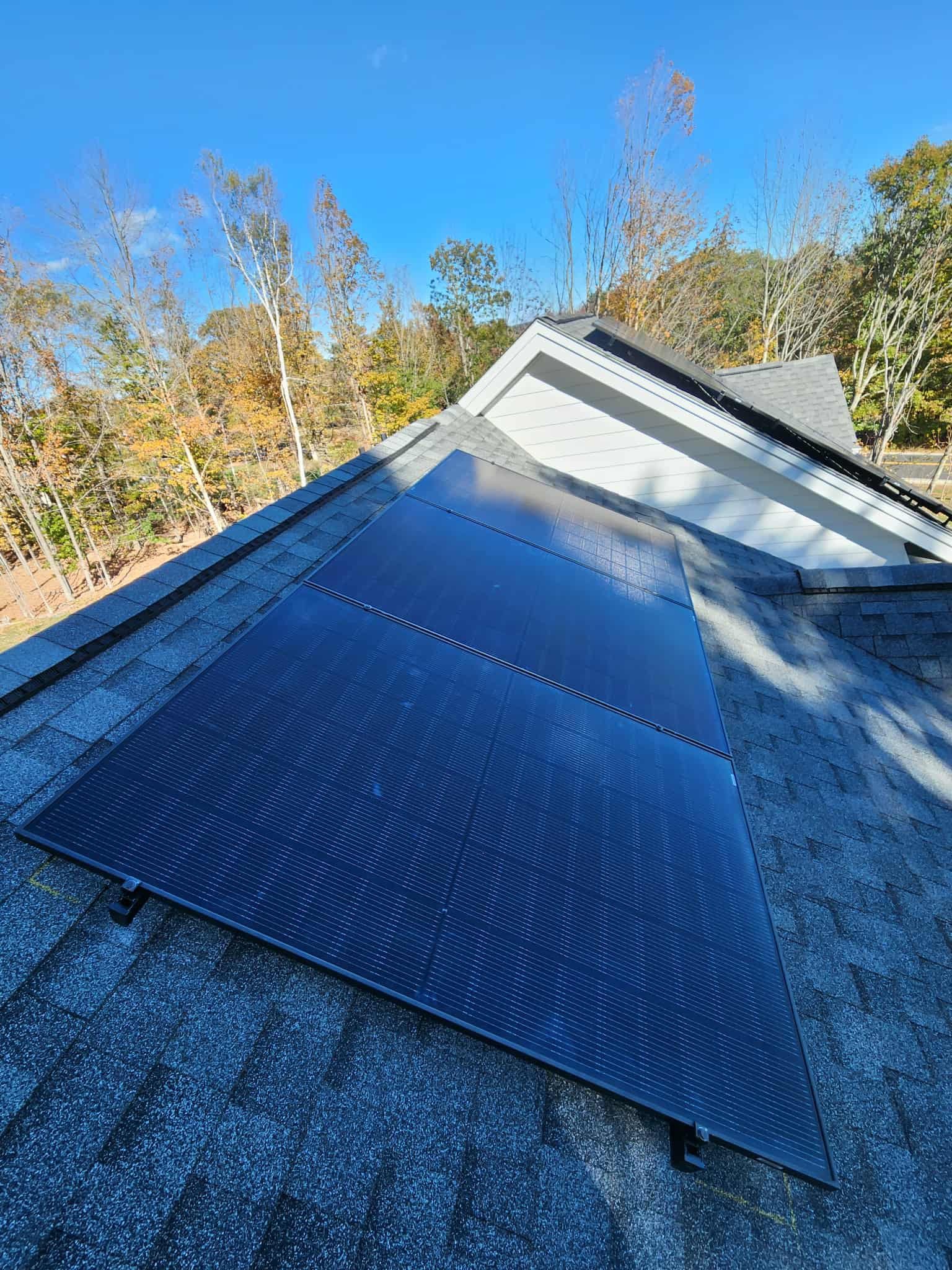 View of a rooftop with solar panels installed on dark shingles, with autumn trees and a blue sky in the background.