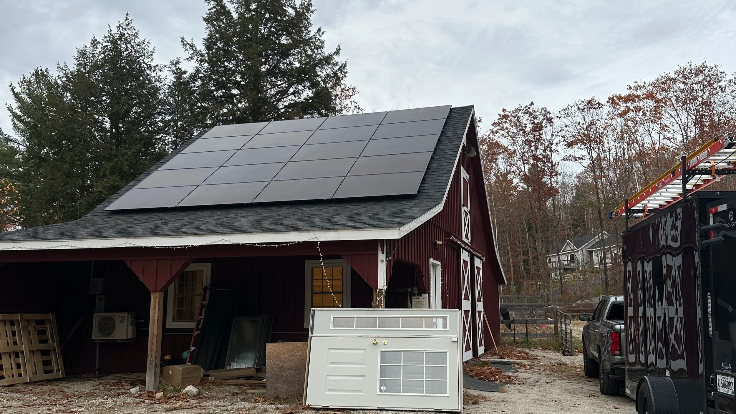 A red barn-style building with solar panels on the roof, surrounded by trees with fall foliage, a truck, and construction equipment in the yard.