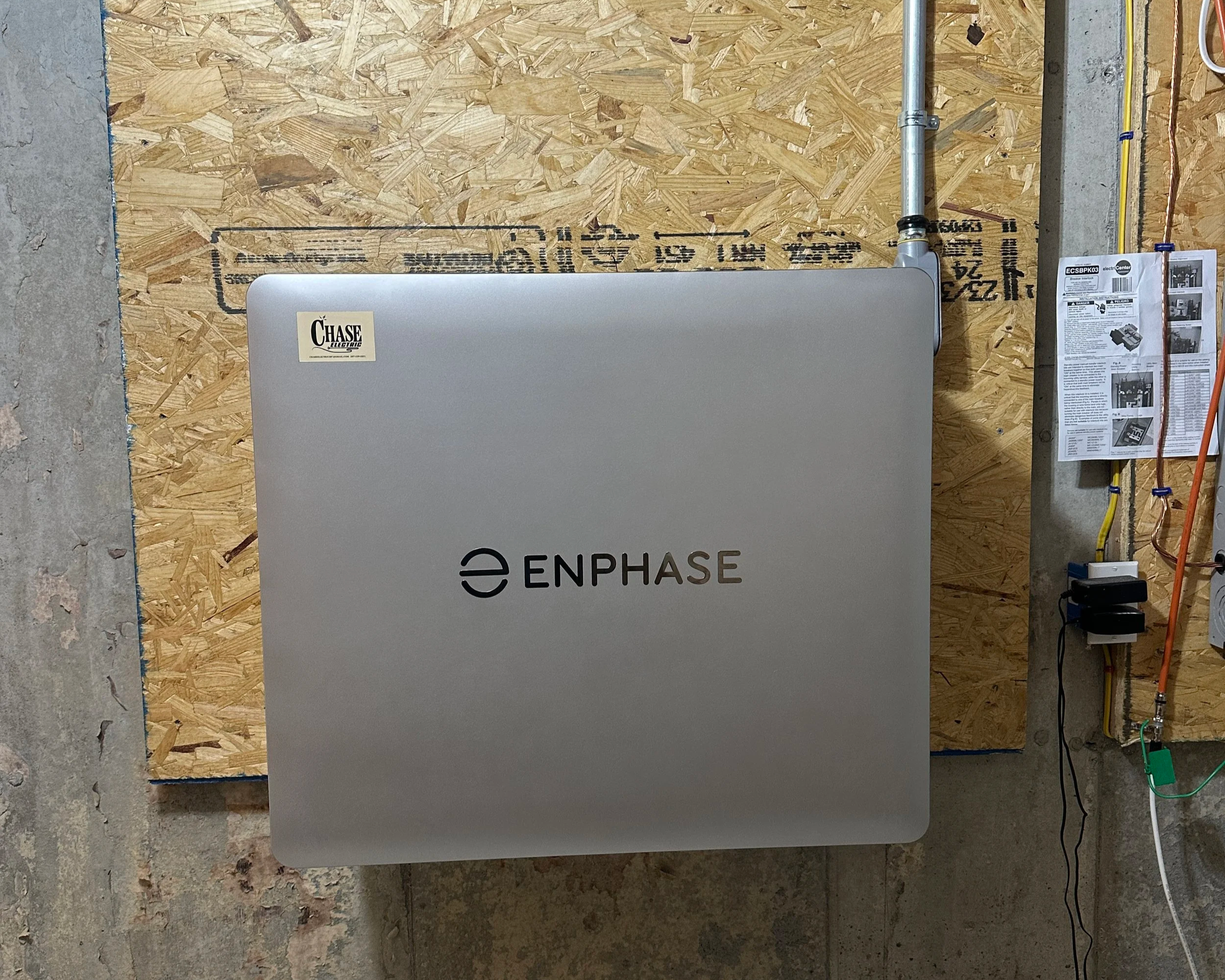 A silver Enphase solar inverter mounted on a plywood wall with a Chase sticker on the top left corner and various electrical outlets and cables on the right side.