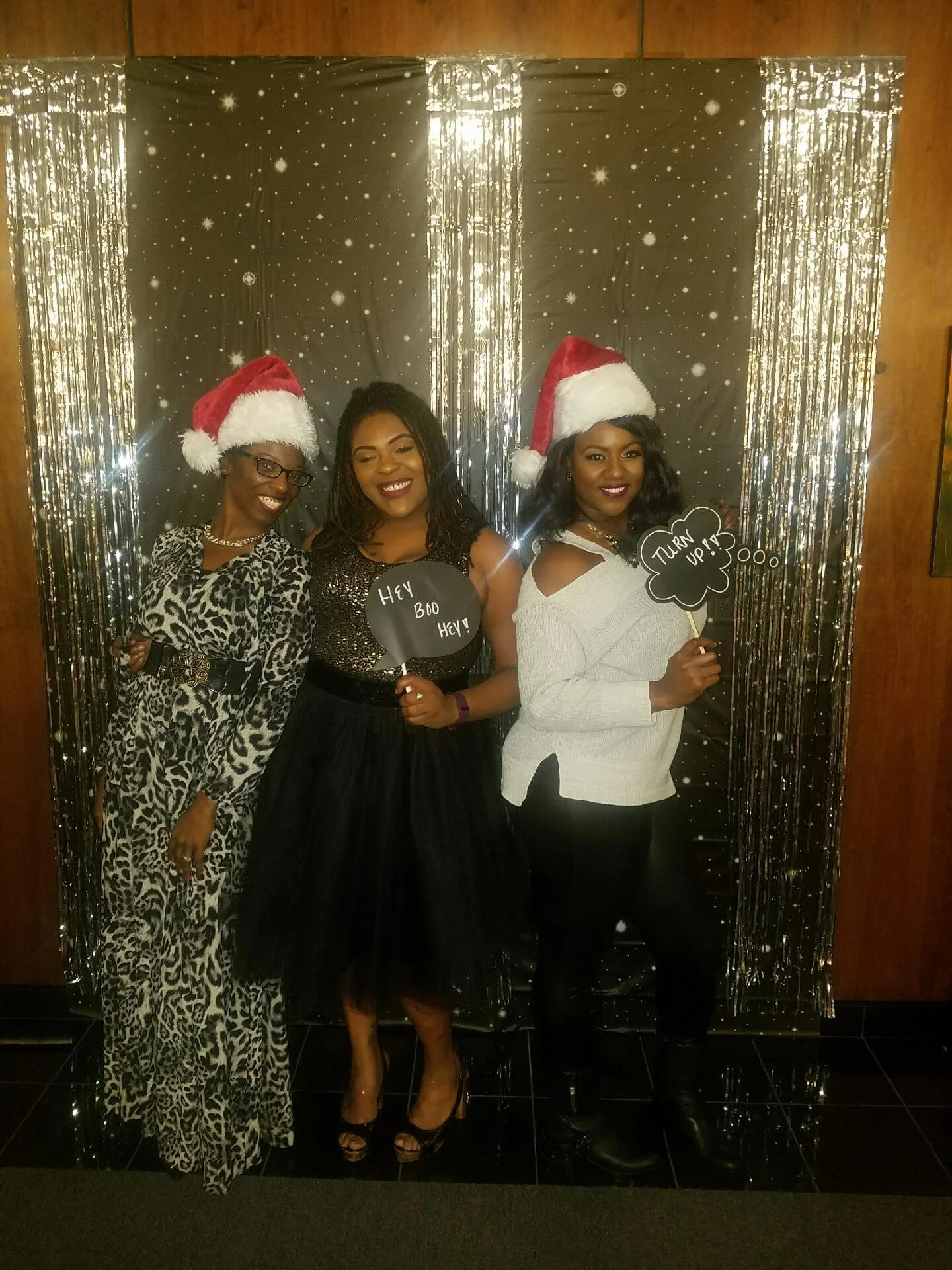 Three women wearing Santa hats, posing together in front of a starry winter themed backdrop with silver tinsel. Two women are holding black speech bubble signs with holiday messages, one says 'Hey Boo Hey!' and the other 'Turn Up!'