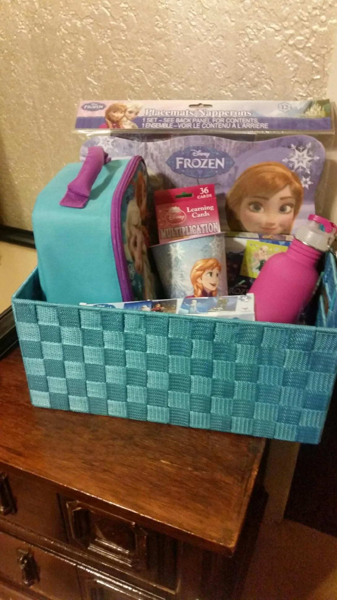 A blue woven storage basket containing Frozen themed items, including a backpack, a water bottle, and a pack of learning cards, placed on a wooden surface