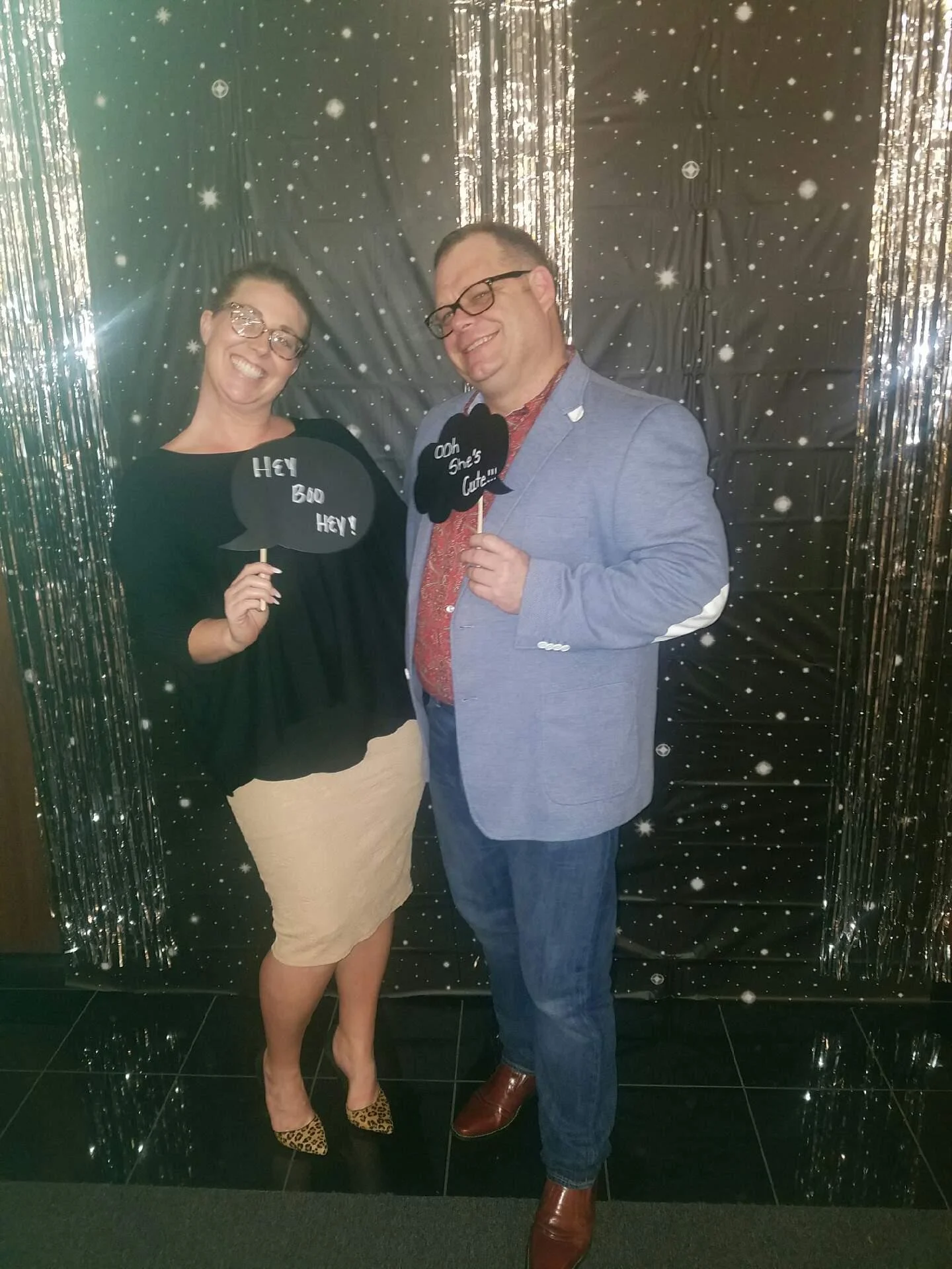 A woman and a man at a party holding speech bubble photo props, smiling, with a black backdrop and silver fringe decorations, giving a festive atmosphere.