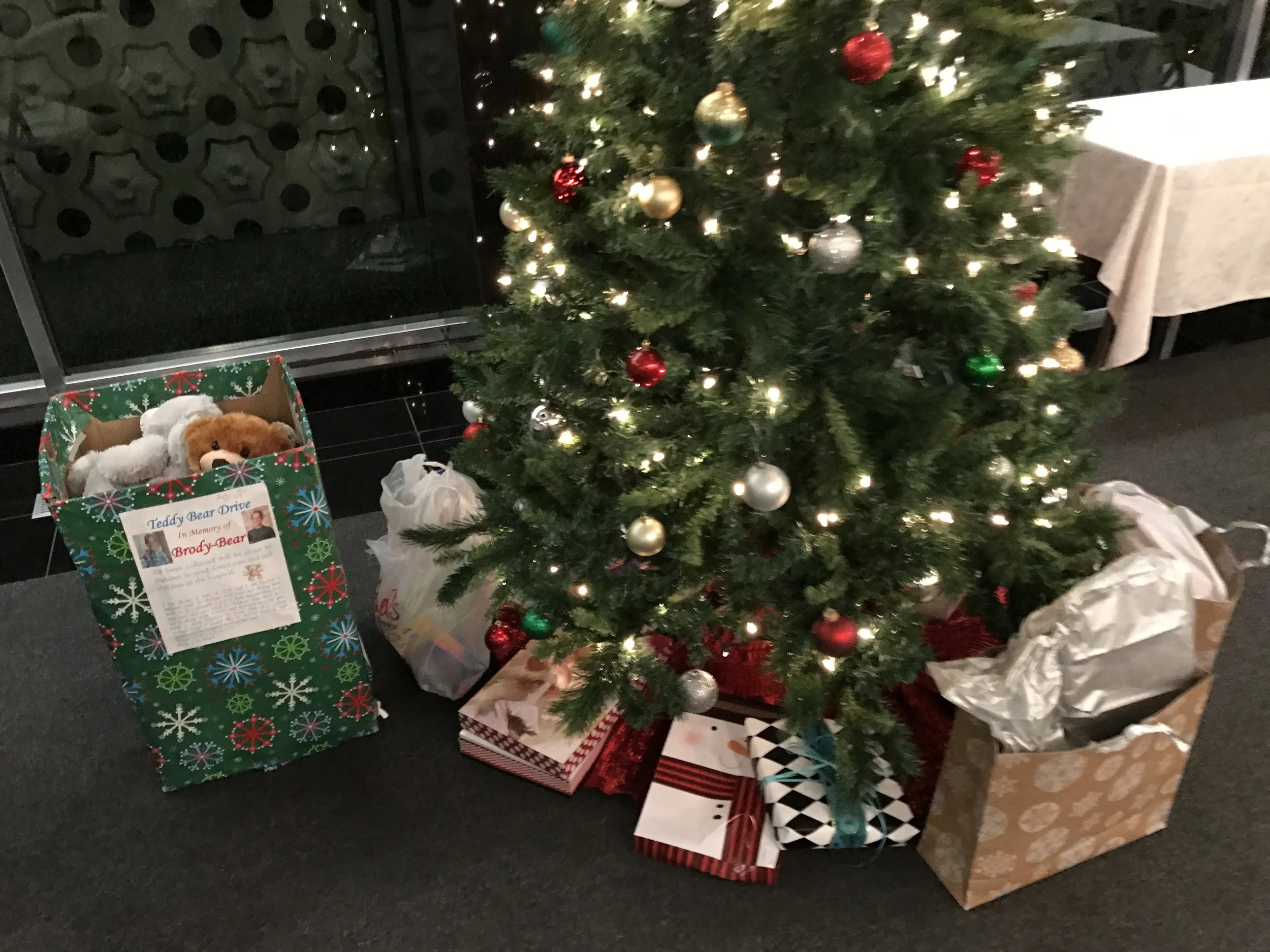 Christmas tree decorated with red, gold, green, and silver ornaments, surrounded by wrapped gifts and gift bags on a dark floor next to a cabinet with a door and a table with a white cloth.