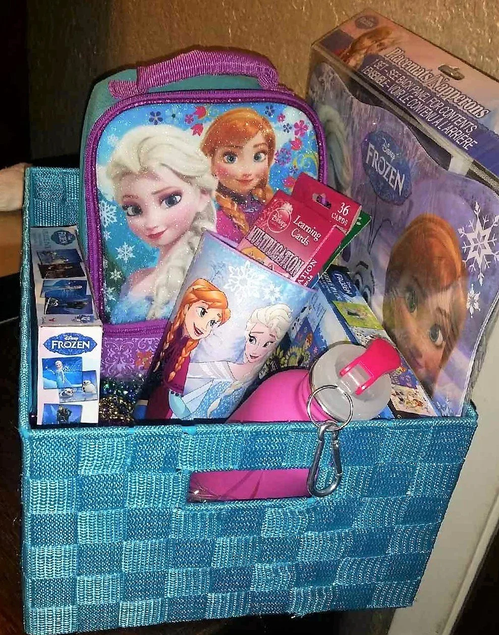 A blue woven storage bin filled with Disney Frozen and Frozen 2 themed items, including a backpack featuring Elsa and Anna, a Frozen puzzle, a water bottle, playing cards, and books.