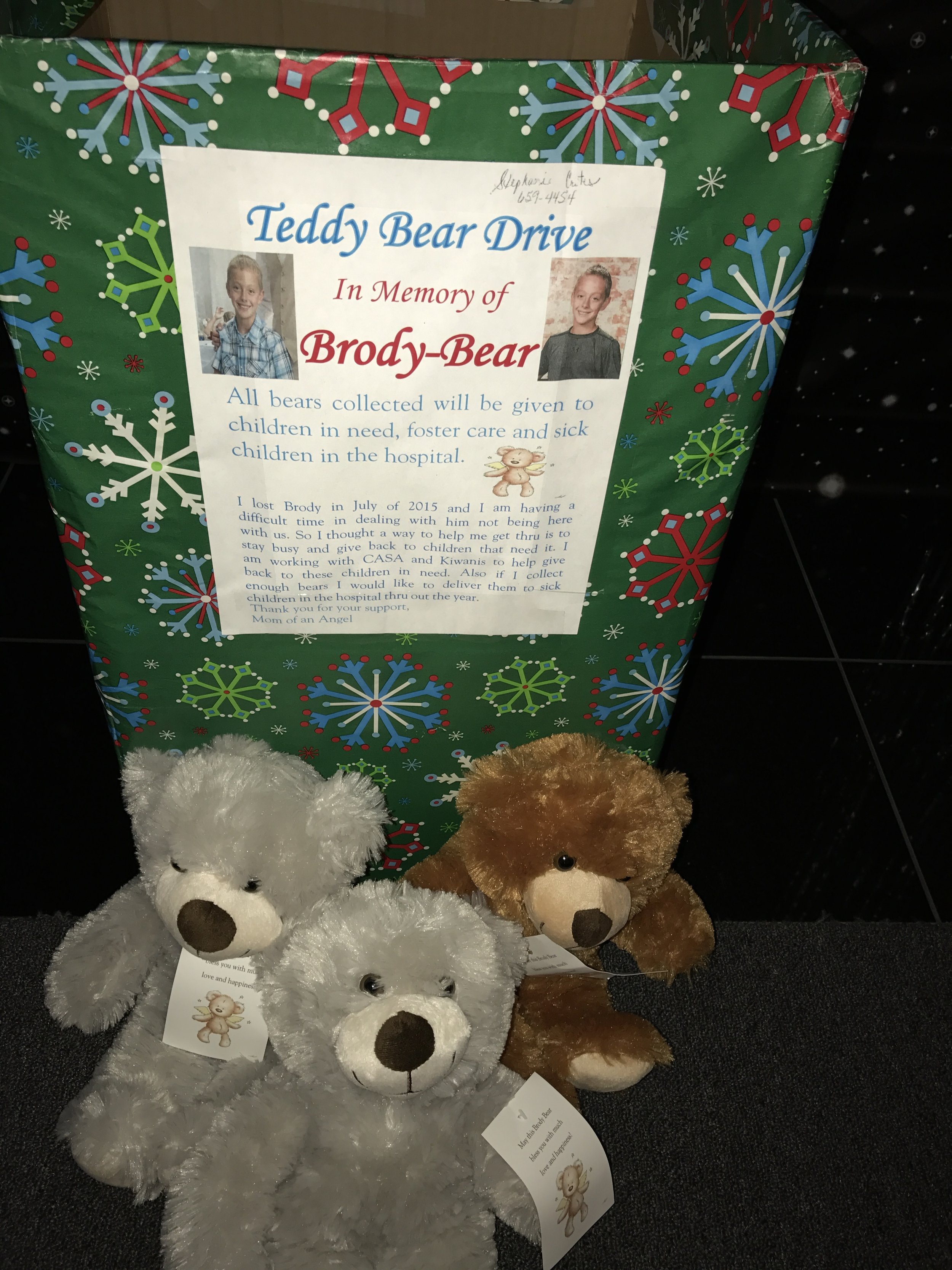 A green Christmas-themed donation box with snowflakes and a photo of two children, in memory of Brody-Bear, for collecting teddy bears for children in need, foster care, and hospitals. In front of the box are three teddy bears, two white and one brow