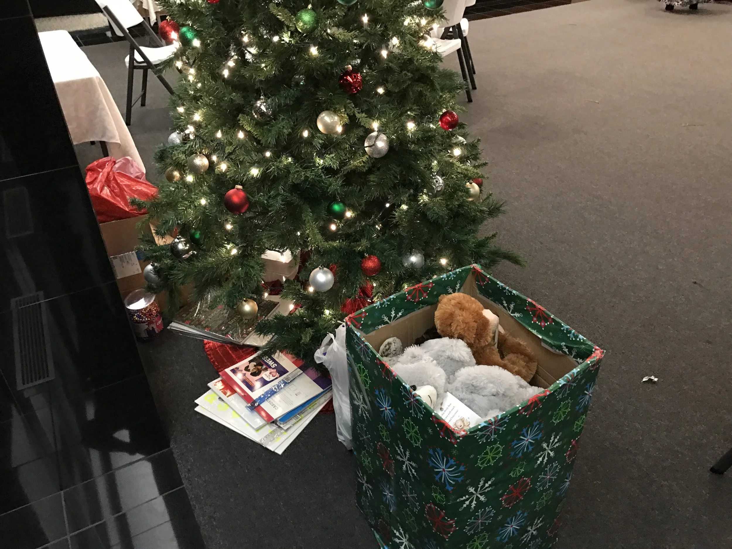 A decorated Christmas tree with ornaments and lights, next to a box of fluffy stuffed animals on the floor, with Christmas magazines and books underneath.
