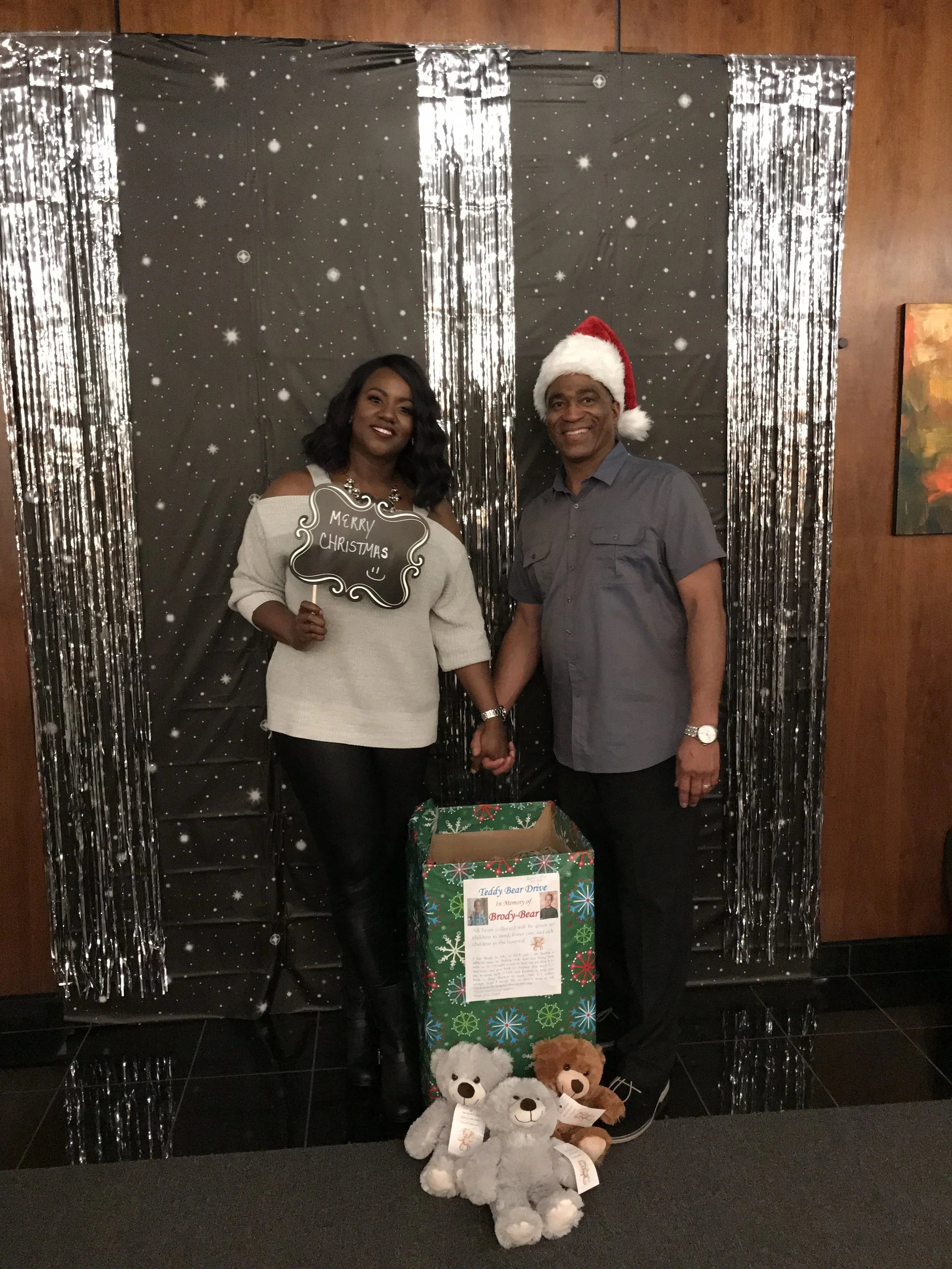 A couple holding hands during a Christmas charity event, with a woman holding a 'Merry Christmas' sign and the man wearing a Santa hat. They stand next to a large gift bag filled with teddy bears and a sign explaining the teddy bear drive. The backgr