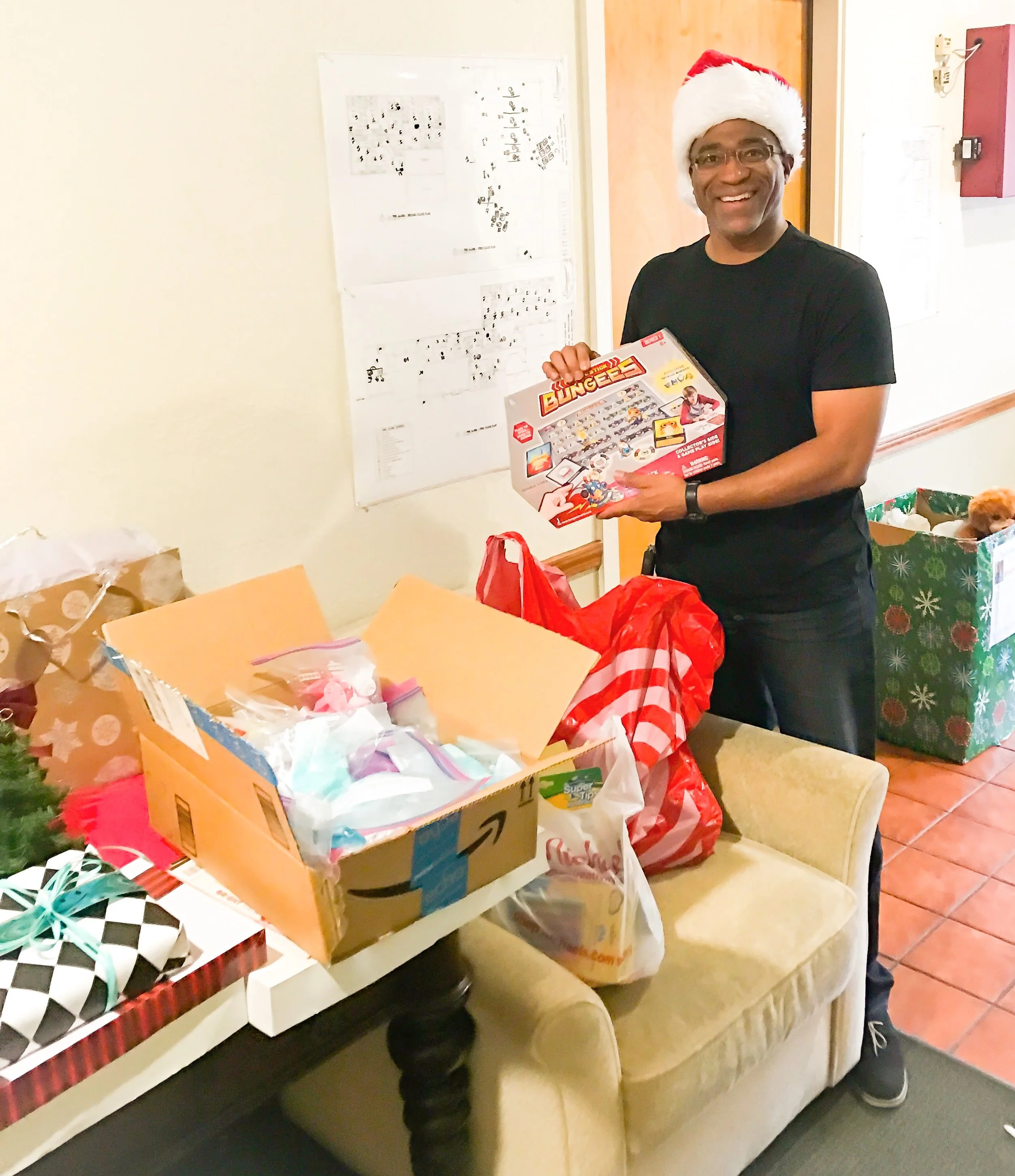 A man wearing a Santa hat is smiling and holding a board game called 'Bungle' at a table with wrapped Christmas gifts and open boxes of presents.