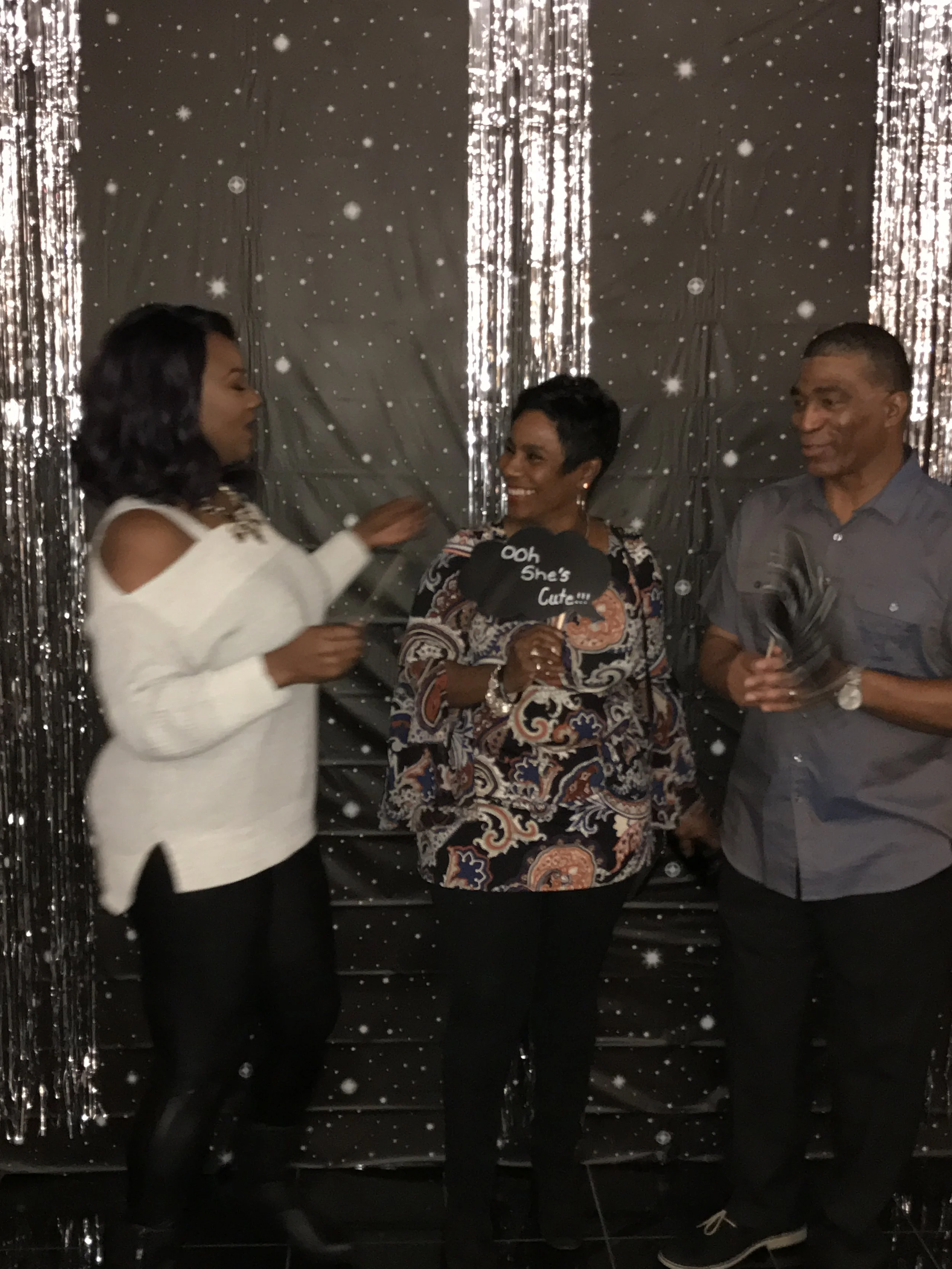 Three people standing in front of a decorative backdrop with silver streamers and snowflake patterns, engaged in conversation at what appears to be a celebration or party.