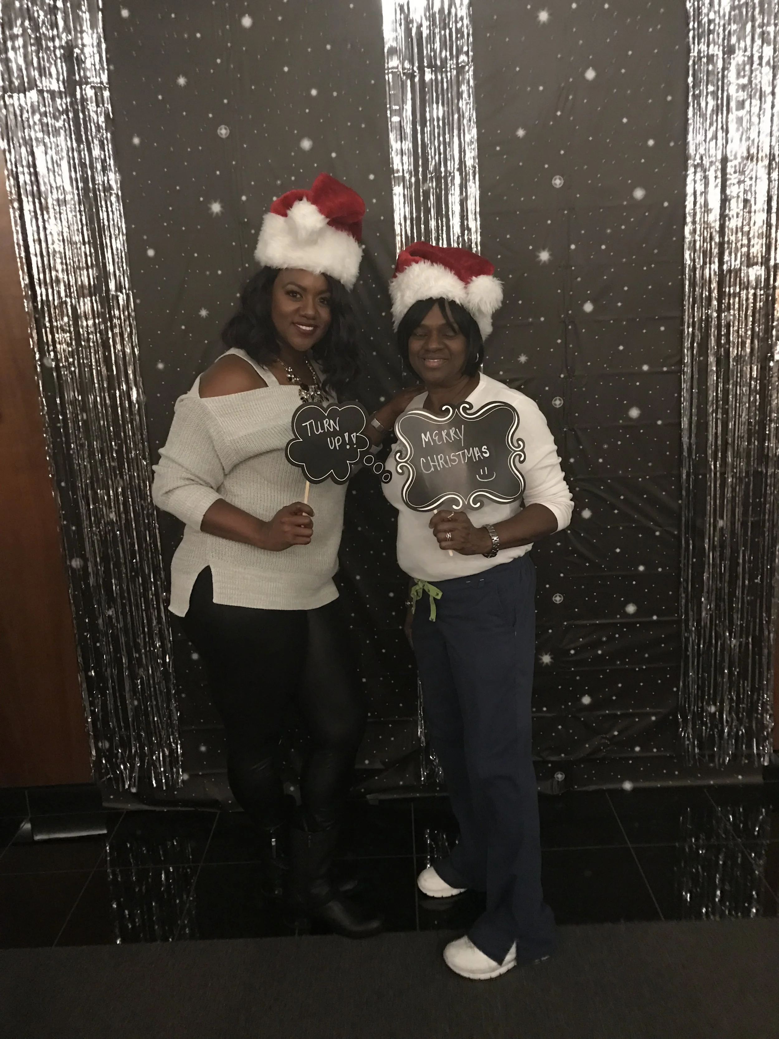 Two women wearing Santa hats standing in front of a black background decorated with snowflakes and silver streamers, holding signs with holiday messages.
