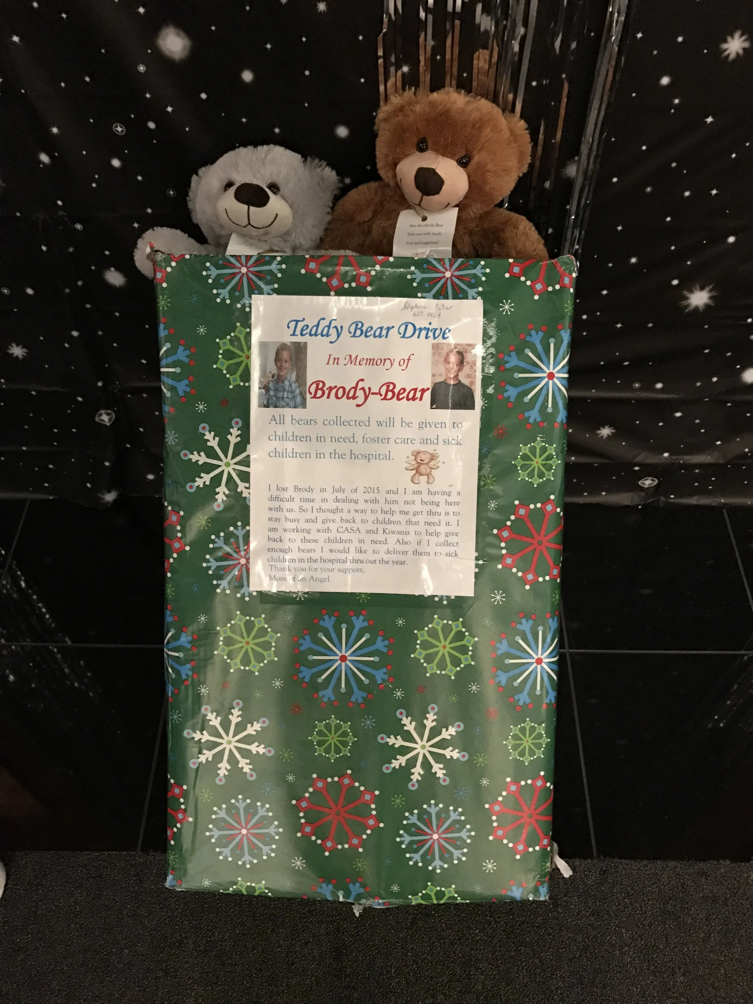 A Christmas donation box decorated with snowflakes, with two teddy bears on top, one grey and one brown, set against a black background with stars.