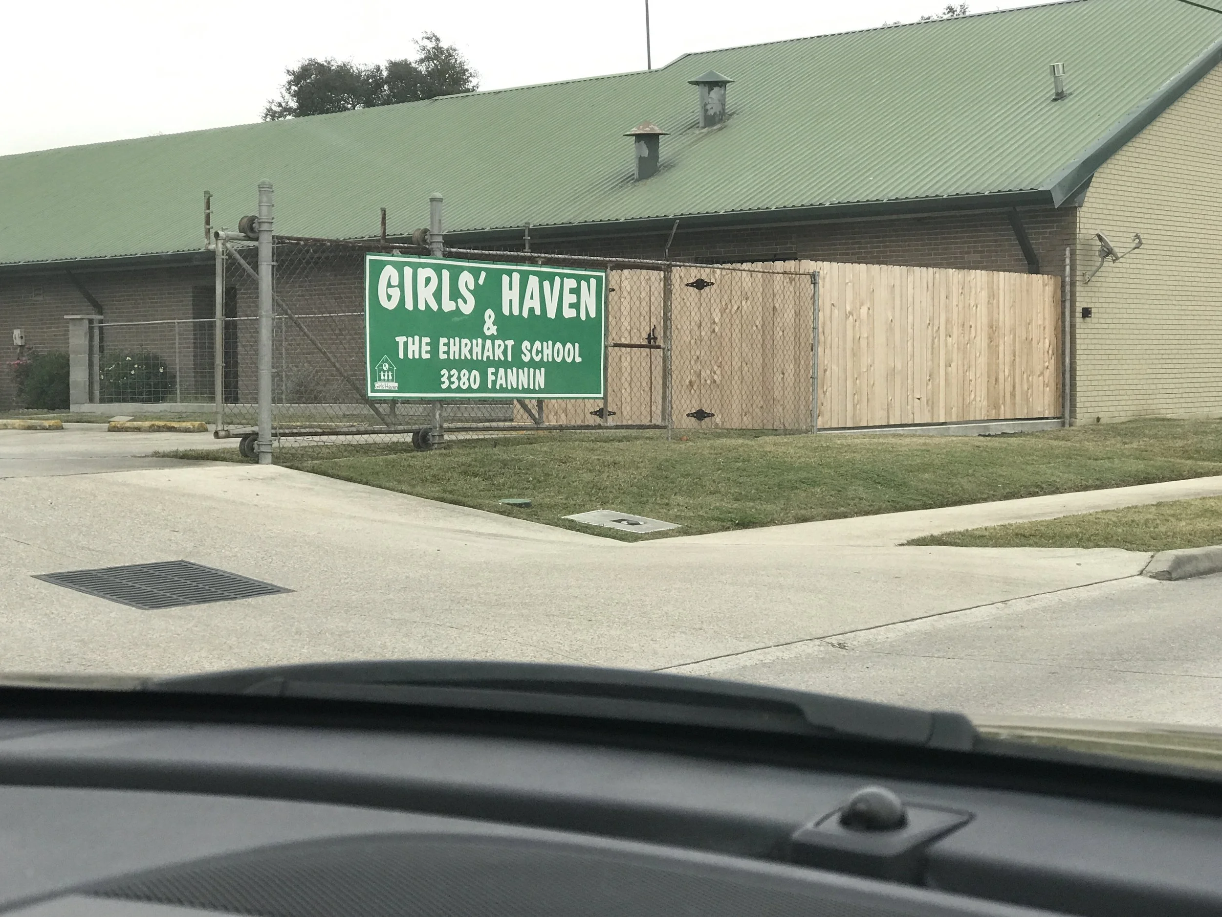 A sign reads Girls' Haven & The Ehrhart School, 3380 Fannin, on a green background, attached to a chain-link fence in front of a building with a green metal roof and beige brick walls.