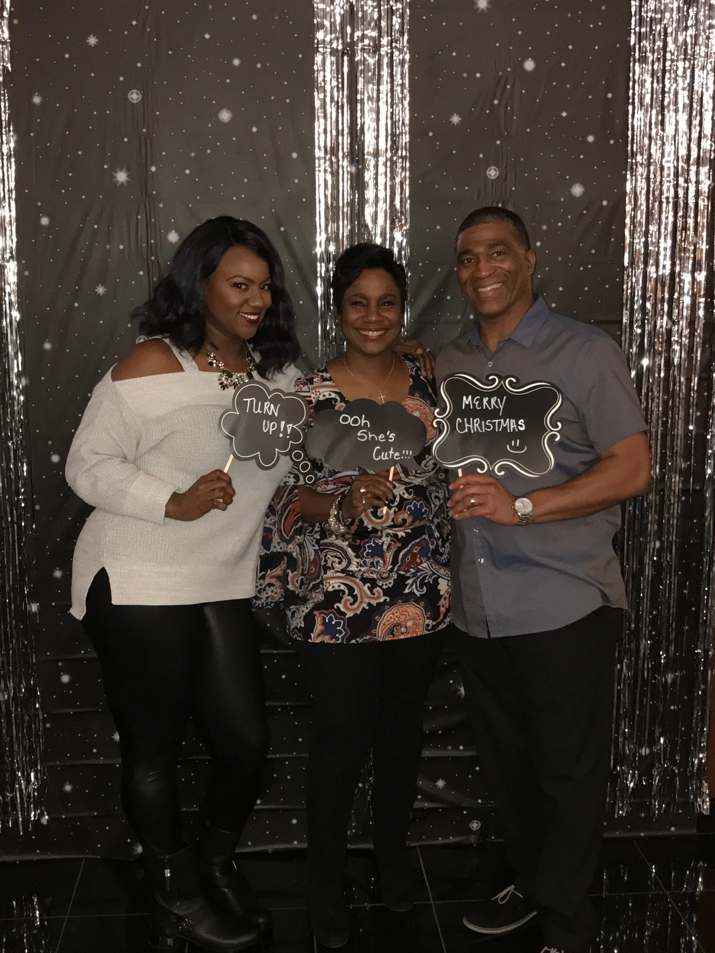 Three adults posing in front of a dark backdrop with silver streamers and snowflake decorations, holding signs with Christmas and holiday messages.