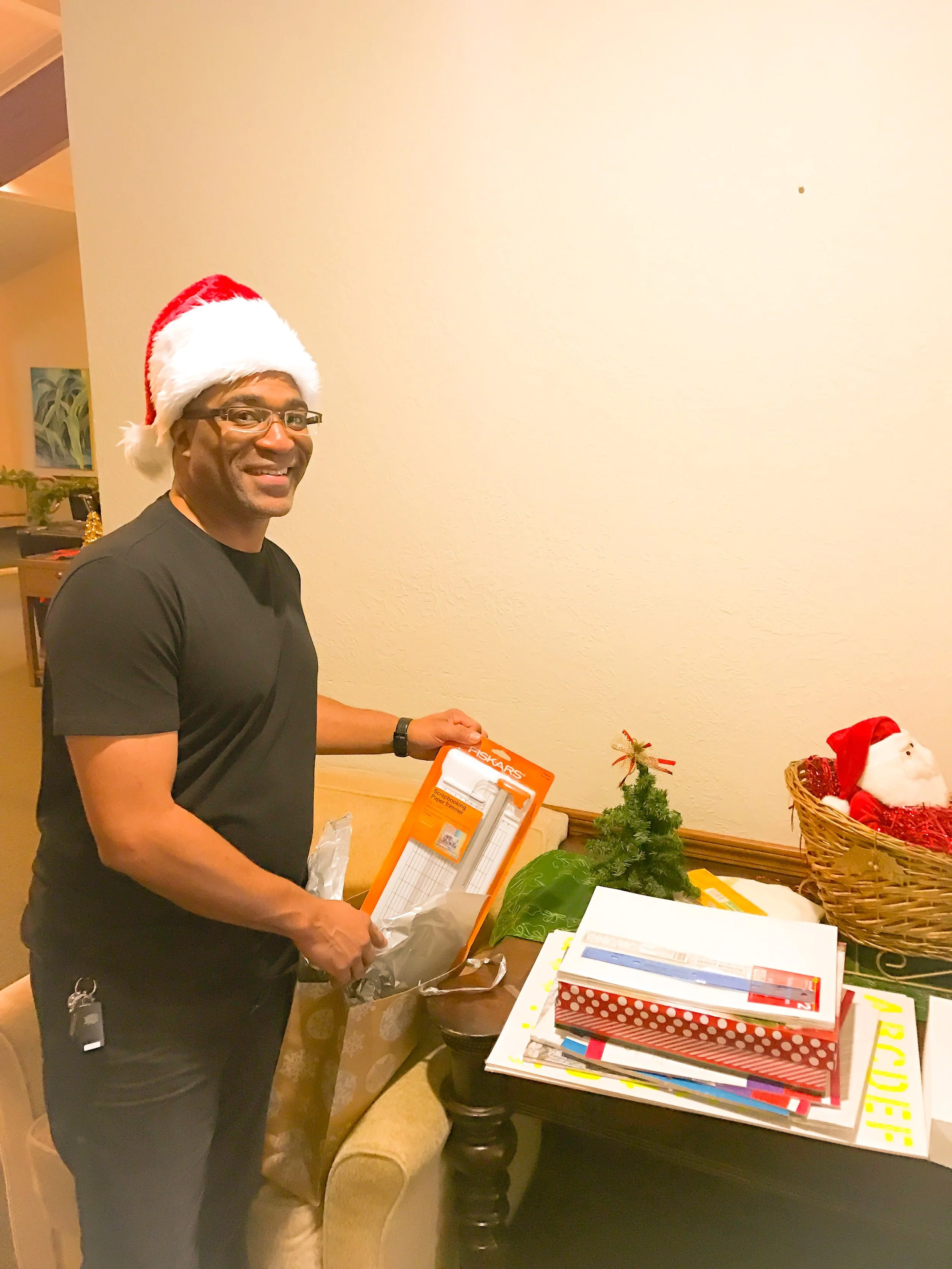A man wearing a Santa hat and glasses is smiling and holding gift wrapping supplies next to a small decorated Christmas tree and a basket with a Santa plush toy. There are holiday gifts and wrapping paper on the table.