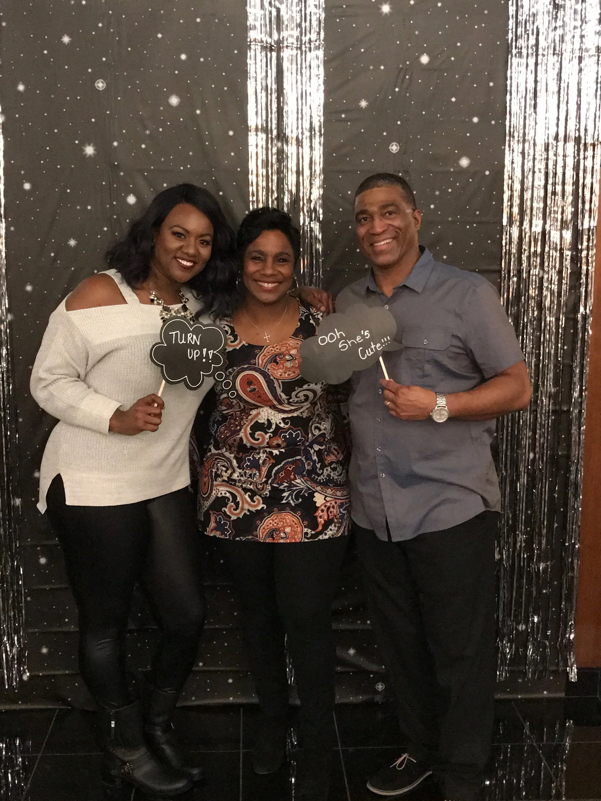 Three people standing in front of a starry night themed backdrop with silver streamers, holding speech bubble signs that say "TURN UP!", "Ooh she's cute!", and "soh she's cute!"