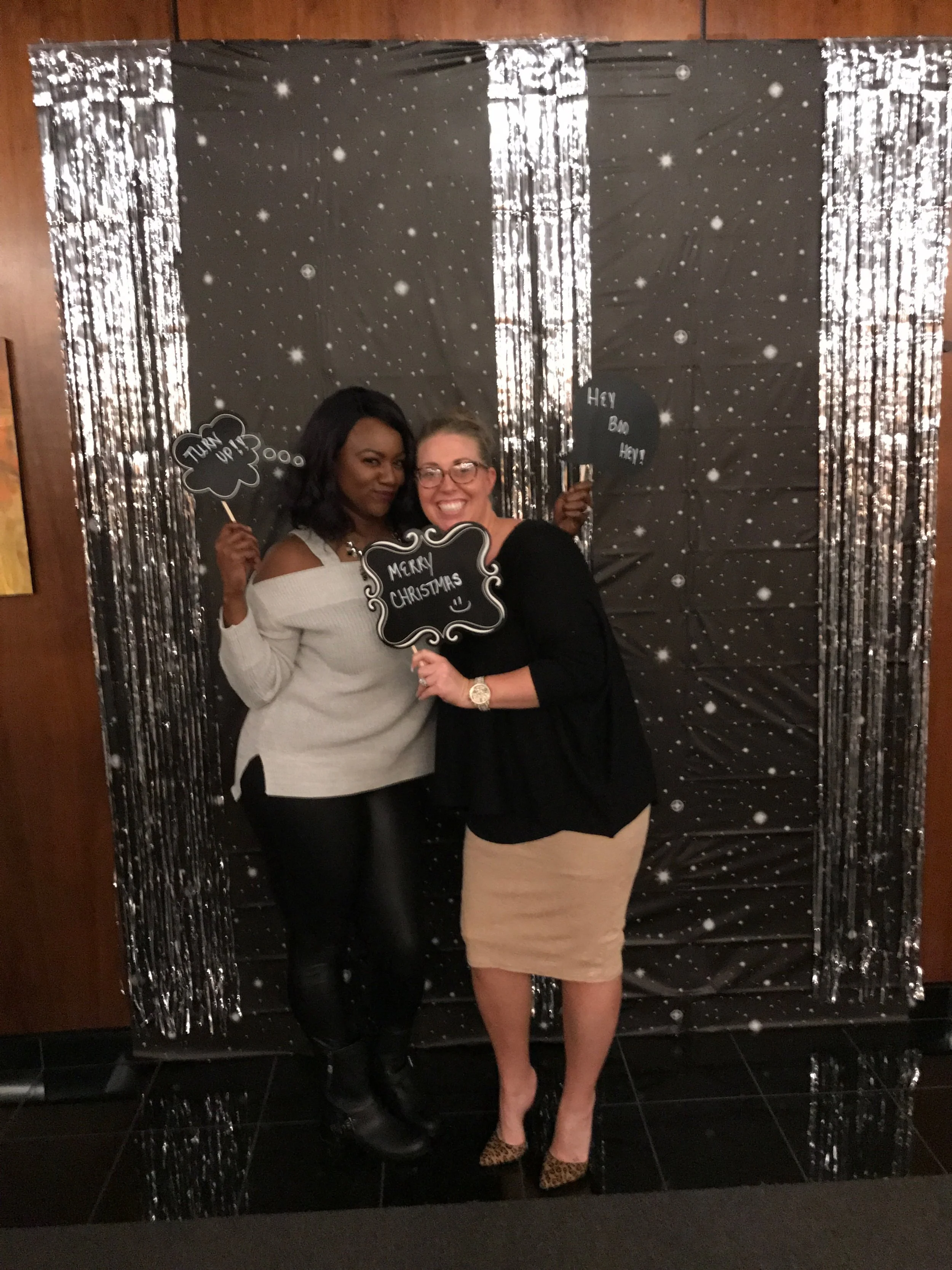 Two women are standing in front of a black backdrop with silver streamers, holding champagne prop signs and smiling. One woman is wearing a white off-the-shoulder top and black pants, and the other is wearing a black top and beige skirt.