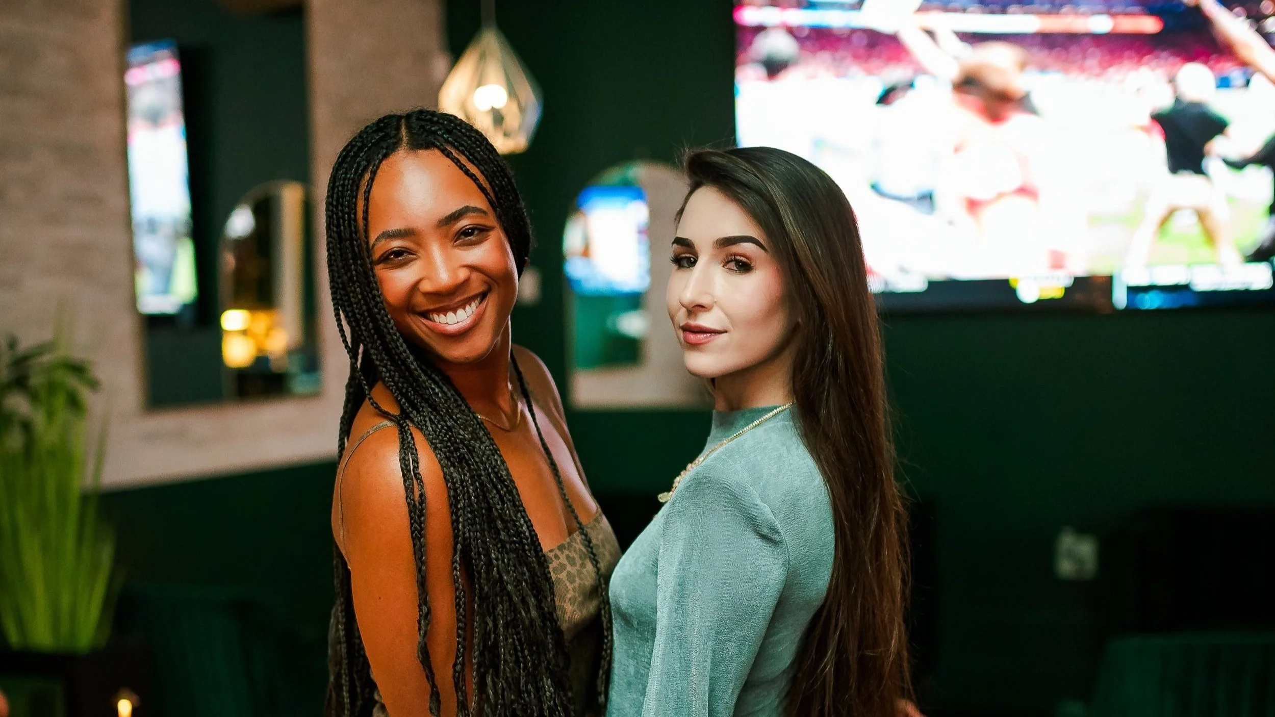 Two women smiling indoors with blurred TV screens in the background.