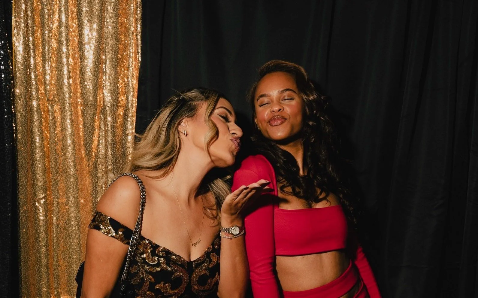 Two women posing in front of a black and gold sequin backdrop, with one in a black and gold dress blowing a kiss toward the other in a red outfit, both smiling.